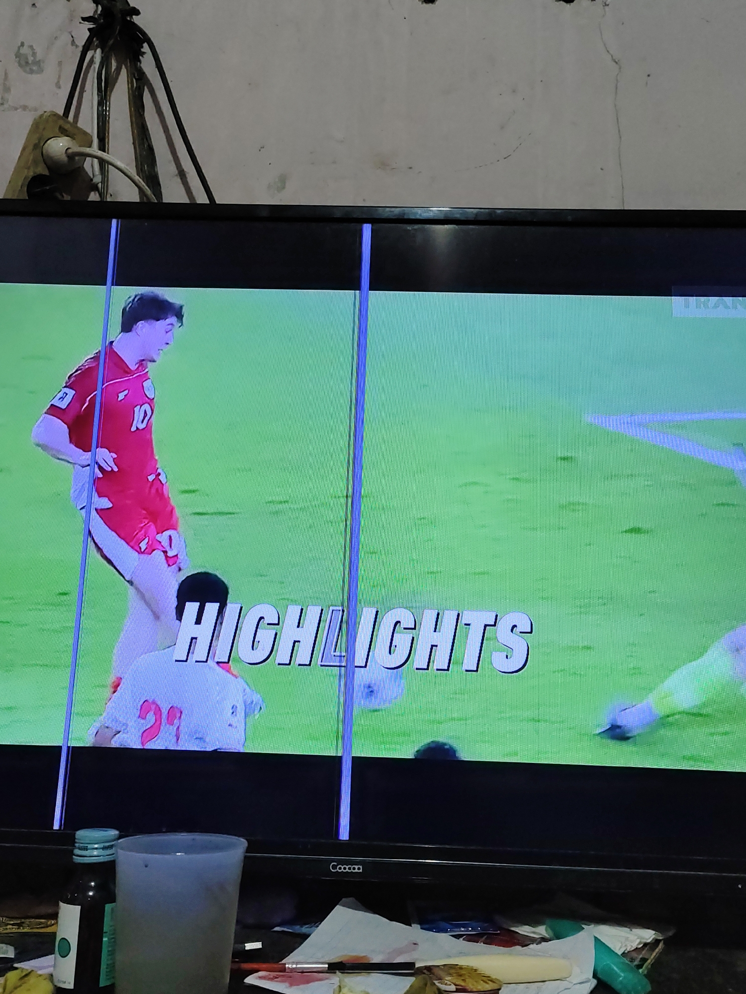 The image depicts a television screen displaying a soccer match highlights. The screen shows two male soccer players on a green field. One player, wearing a red jersey with the number 10, is positioned in the foreground, facing to the right. Another player, partially visible and wearing a white jersey with the number 21, is kneeling in front of him. The text "HIGHLIGHTS" is overlaid on the screen in large, white, 3D letters. The television screen itself is showing vertical lines of distortion. The television is placed on a black stand, and below the screen, there are various objects including a bottle with a blue cap, a translucent cup, and some papers. The background behind the television appears to be a light-colored wall with exposed electrical wires and an electrical outlet. The image is taken from a slightly low angle, looking up at the television. The scene is indoors, likely in a residential setting, and is not indicative of a specific location within South Jakarta, Indonesia.