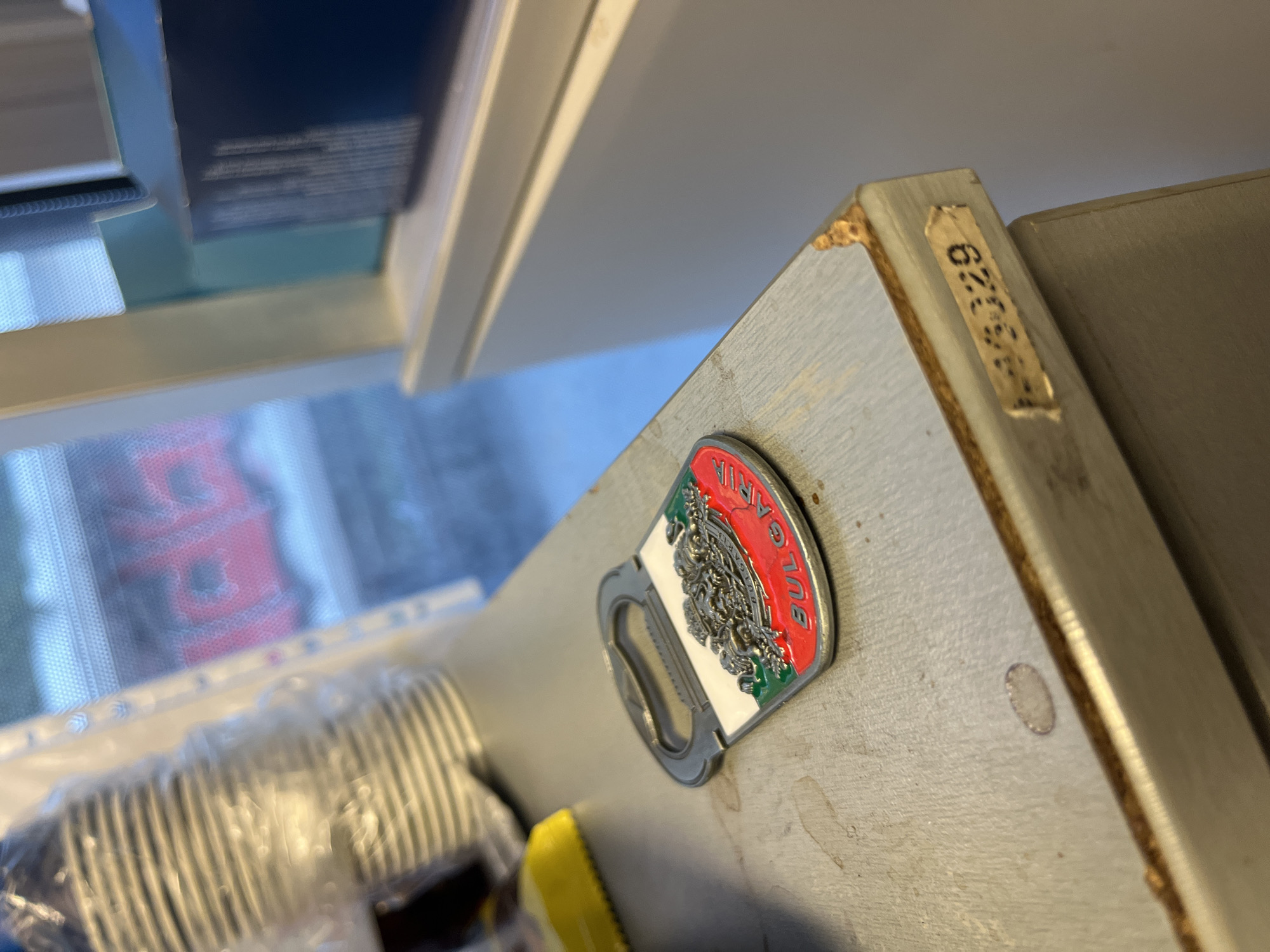 A close-up, eye-level shot focuses on a metallic bottle opener with a design depicting the Bulgarian coat of arms and the word "BULGARIA." The opener is positioned on a light gray, textured surface, likely a countertop or shelf, which shows some signs of wear and tear, including a chipped corner with a sticker that has the text "L-430329". In the background, out of focus, are stacked white paper cups inside a clear plastic wrapper, a dark blue box, and a window showing a blurred view of a red object, possibly a vehicle or building. The overall environment appears to be indoors, with diffused light. The scene is devoid of any people or immediate action. The location is not specified in the image, and therefore cannot be determined to be the city of Varna, Bulgaria.