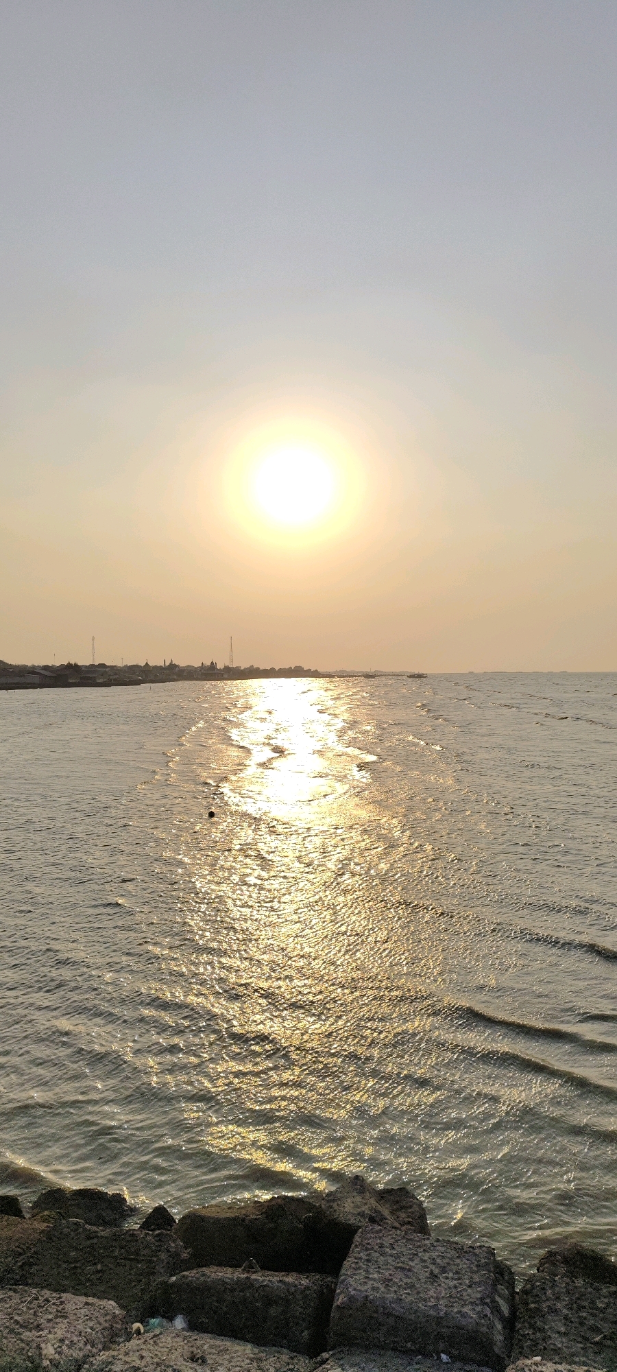 The image captures a serene sunset scene at the coast of Tasikagung, Indonesia. The sun, a bright, luminous orb, hangs high in the hazy sky, casting a golden path across the rippling water. In the distance, a low-lying coastline is visible, dotted with buildings and what appear to be telecommunication towers, suggesting a developed settlement. The foreground is occupied by a rocky shoreline, with large, dark stones forming a natural barrier. The overall atmosphere is one of tranquility and natural beauty, accentuated by the warm glow of the setting sun reflecting on the sea.