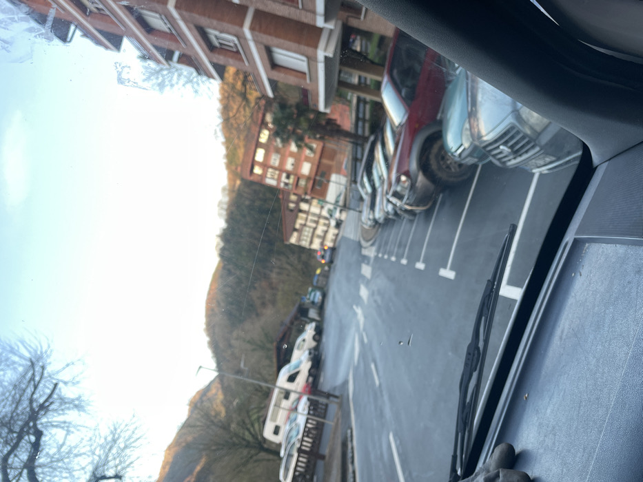 The image presents a daytime street view in Zestoa, Spain, observed from within a vehicle, with a dark dashboard and a windshield wiper visible in the immediate foreground. The scene depicts a paved road with white line markings, including dashed lines, solid lines, and pedestrian crosswalks. To the right, a series of vehicles is parked in designated street parking spaces marked with white 'T' symbols; these include a light-colored SUV, a dark red SUV with off-road tires, and several other cars receding into the distance. To the left of the road, a white camper van, a white delivery van, and other cars are parked. The background features multi-story residential buildings; a prominent brick-red building with multiple balconies stands on the right, while other lighter-colored structures are visible further down the street. Beyond the buildings and road, tree-covered hills or mountains rise under a clear, bright sky. Bare-branched trees are present in the left foreground and on the distant hills. A street lamp is visible on the left side of the road. No individuals are present in the depicted scene.