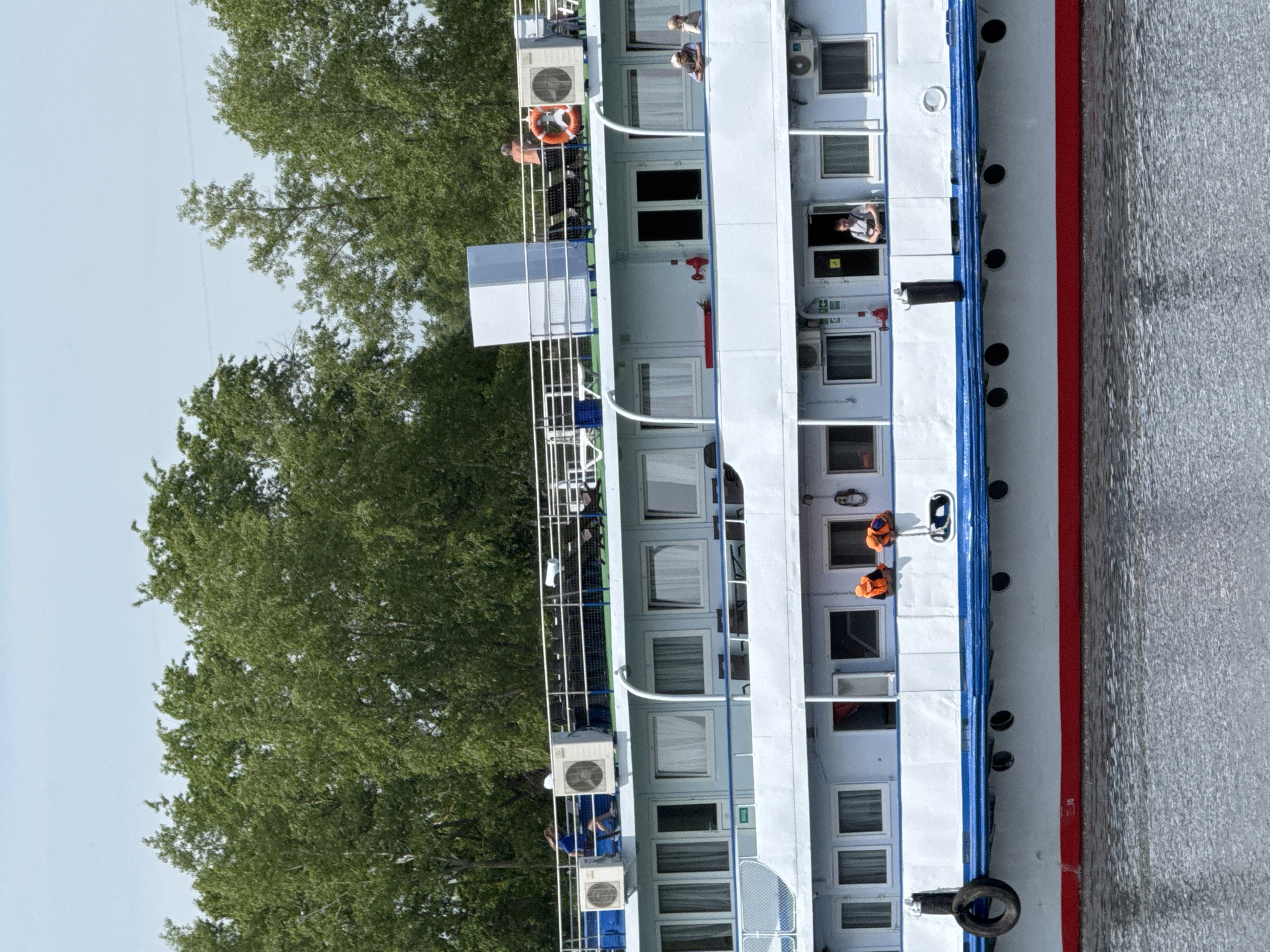 The image depicts a multi-level river cruise ship. The ship is white with blue and red trim. There are numerous windows and at least one person visible in a window, potentially staff. People are on the upper deck of the ship. A few people are wearing bright orange life vests. The ship is adjacent to a body of water, and a dense group of tall, green trees is visible on what appears to be the riverbank. The image is taken during daylight. The location is potentially Chaikovsky, Russia.