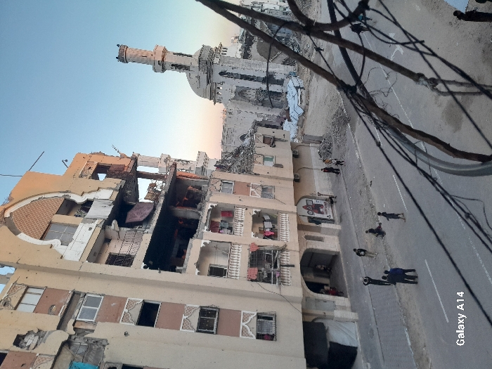 A war-torn building with significant damage and missing walls stands beside a street with people walking. Smoke and rubble are visible within the damaged structure. In the background, a mosque with a minaret is partially destroyed. Power lines stretch across the top of the image. The overall scene depicts devastation and potential conflict in an urban setting.