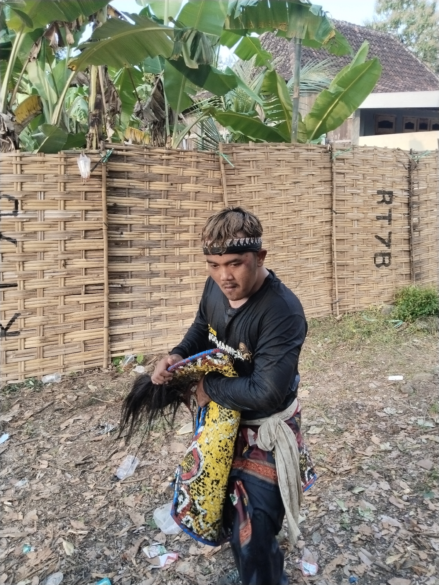 The image captures a man standing in an outdoor environment in Jenggrik, Indonesia. He is attired in a long-sleeved black shirt, a patterned sarong around his waist, and a patterned headband. The man is holding a traditional Indonesian horse dance prop known as a "Kuda Lumping," distinguished by its horsehair tail and yellow and black coloration. The ground is covered in leaves and litter. Behind him stands a woven bamboo fence, with banana trees and a building visible in the background. The text "RT 7B" is painted on the fence.
