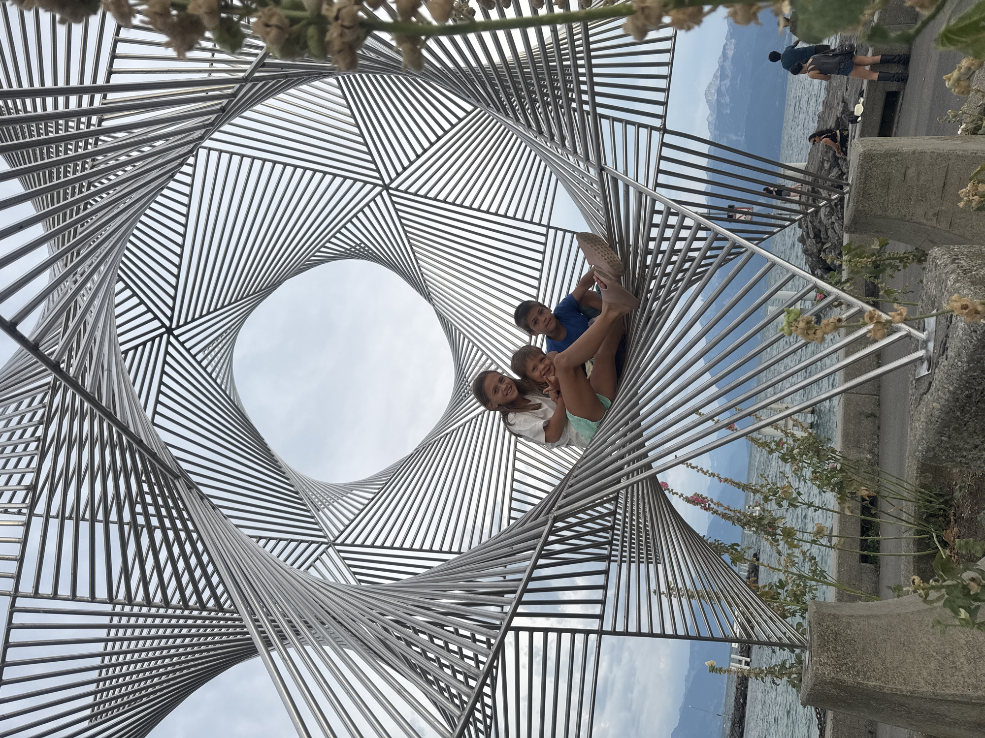 A wide-angle, low-angle shot captures a modern metal sculpture on the shores of Lake Geneva in Lausanne, Switzerland. Three children are seated within the sculpture's geometric, spiraling frame. The oldest child, a girl, is in the center, wearing a white t-shirt and teal shorts, with her arm around a younger child. To her left, a younger girl in a light-colored shirt and denim shorts smiles and leans in. To the girl's right, a boy in a blue t-shirt and dark shorts looks towards the camera, his legs extended. The sculpture is made of numerous parallel metal rods that create a dynamic, twisted effect, allowing views of the sky and the lake beyond. In the background, the calm waters of Lake Geneva are visible, with distant mountains on the horizon. The shoreline features a rocky embankment, a paved walkway, and concrete planters with dry vegetation. Other people are visible further down the promenade, some seated on the rocks and others walking. The sky is overcast, contributing to a soft, diffused light.