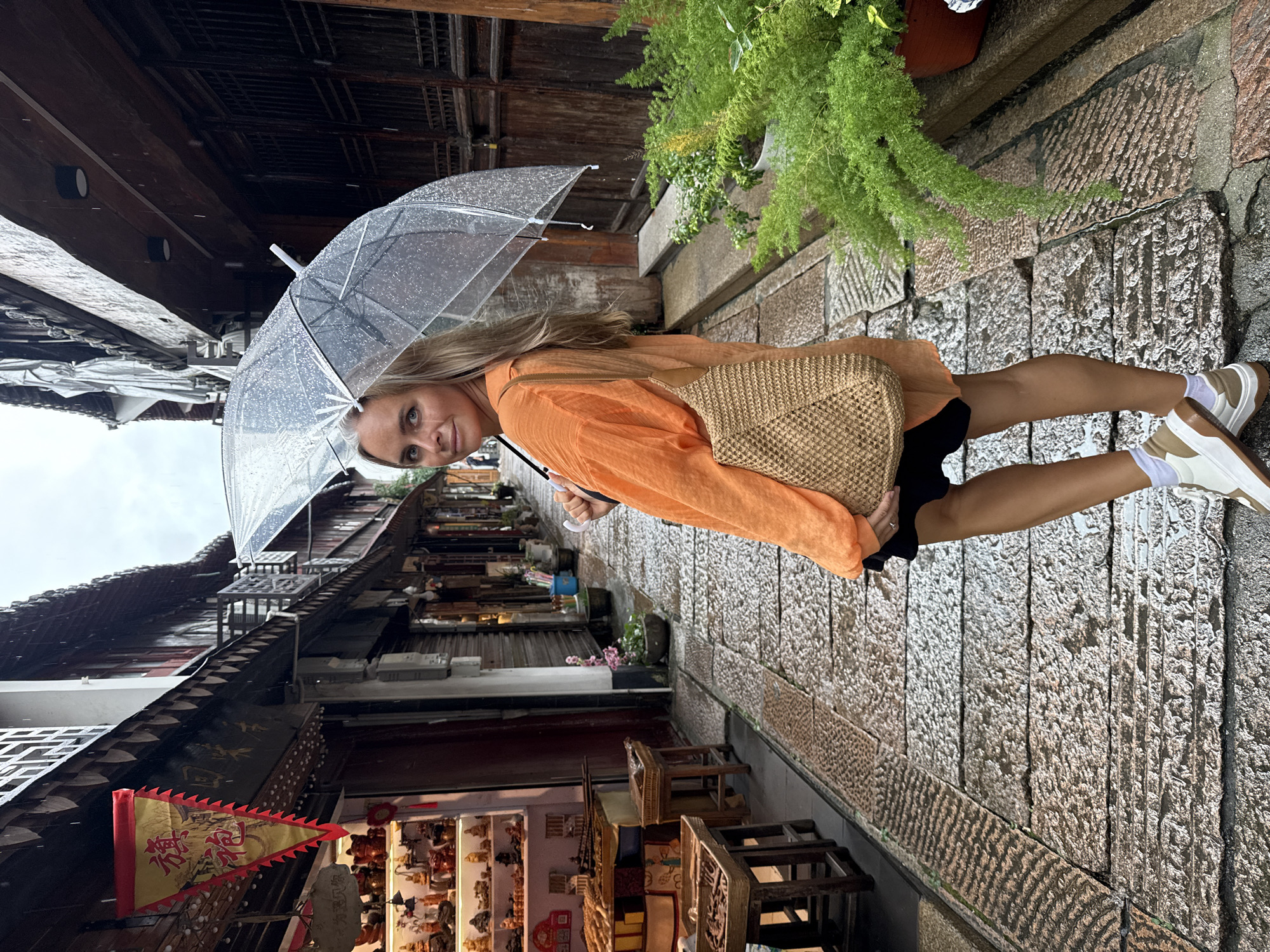 A Caucasian woman stands on a wet, stone-paved street in a traditional Chinese town, holding a clear umbrella over her head. She is facing away from the camera, looking over her left shoulder, with a slight smile. The woman is wearing an orange oversized blouse, a black skirt, and white sneakers with brown accents. She carries a woven straw bag over her right shoulder. The street is lined with old wooden buildings, some with shops displaying goods. A red and yellow banner hangs from one of the buildings on the left. Lush green plants are visible in pots to the right of the woman. The weather appears to be rainy, with visible raindrops on the umbrella and the wet pavement. The scene suggests a picturesque tourist area in Qingpu District, China.