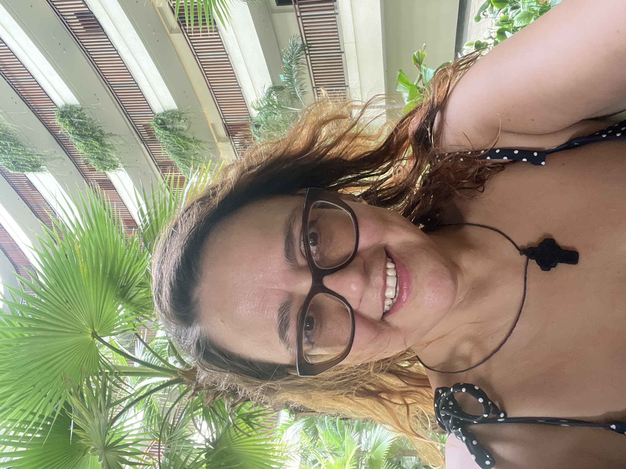 The image is a selfie of a woman with brown, wavy hair and glasses. She is wearing a black and white polka dot top and a black necklace with a cross pendant. She is smiling and looking at the camera. In the background, there is a building with multiple balconies and hanging plants, as well as palm trees. The location is likely a hotel or resort in Nha Trang Ward, Vietnam, suggested by the tropical foliage and the architectural style.