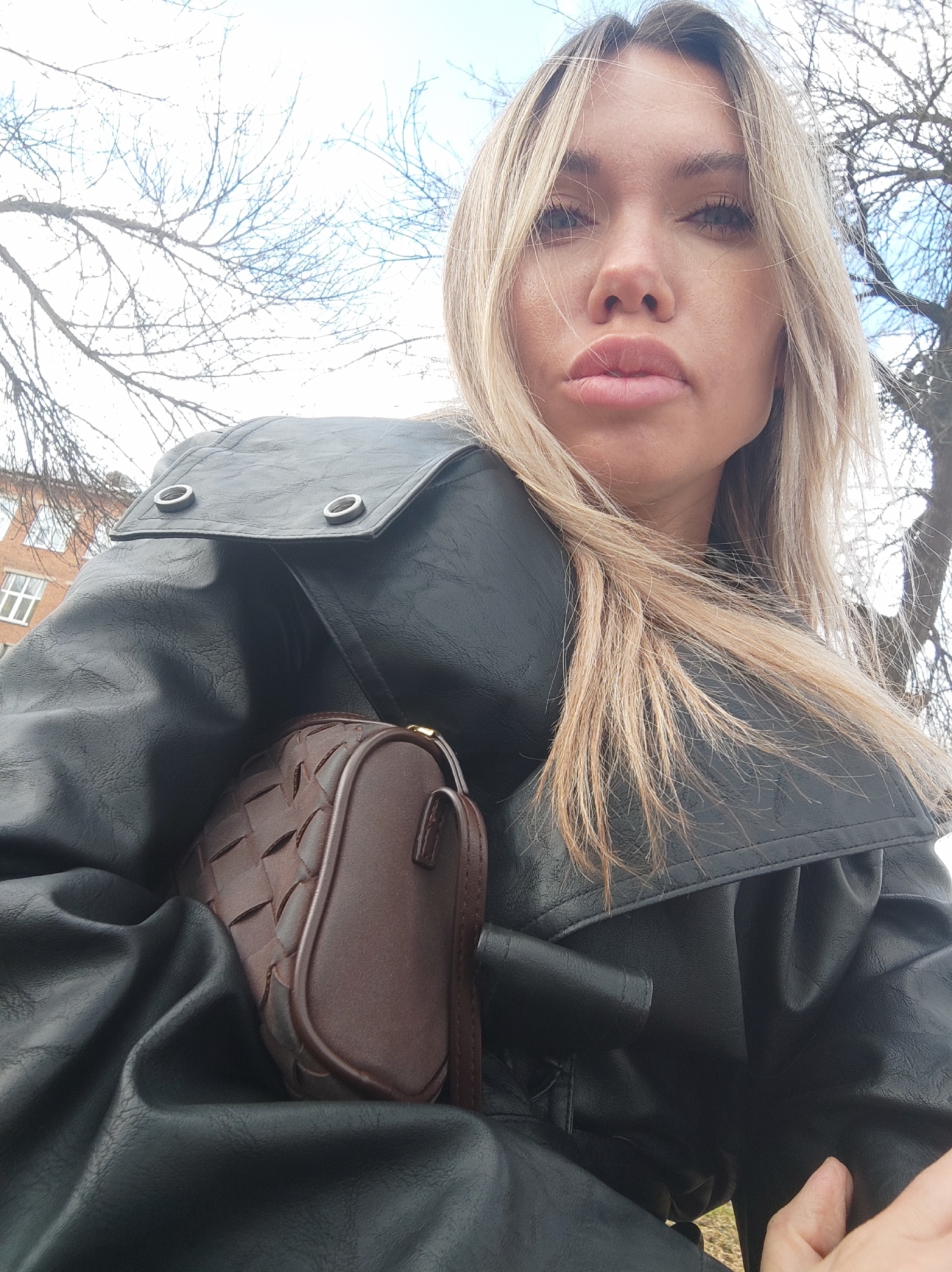 A woman is pictured from a low angle, looking downward. Her hair is blonde and slightly disheveled, some strands falling across her face. She has a neutral expression with slightly pursed lips. She is wearing a black, leather-like jacket with a wide collar, visible buttons and a dark brown woven-texture handbag. The background shows bare tree branches against a bright sky, with a glimpse of a brick building with white-framed windows in the upper left. The picture was taken in Votkinsk, Russia.