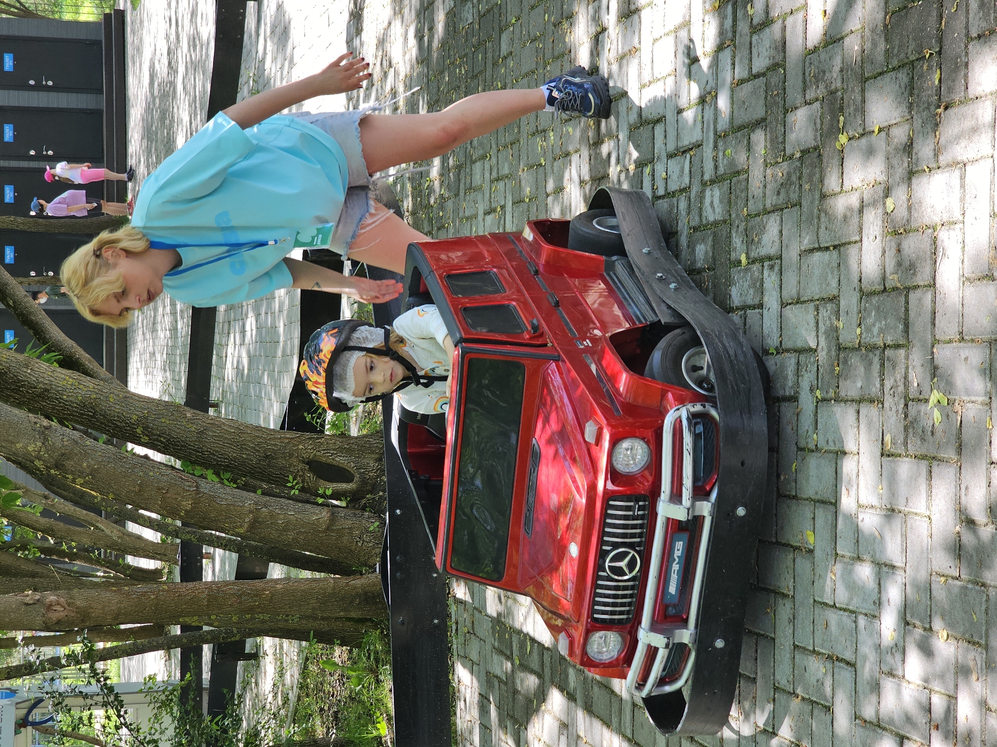 A young child wearing a helmet is seated in a red, toy-sized Mercedes-Benz AMG vehicle. A young woman is standing next to the vehicle, appearing to interact with the child. The scene is set on a paved area with patterned gray bricks, possibly a pedestrian zone. There are tree branches visible in the background. A building and other figures, including people in pink and blue shirts, are in the distant background. The location is likely in Khimki, Russia.