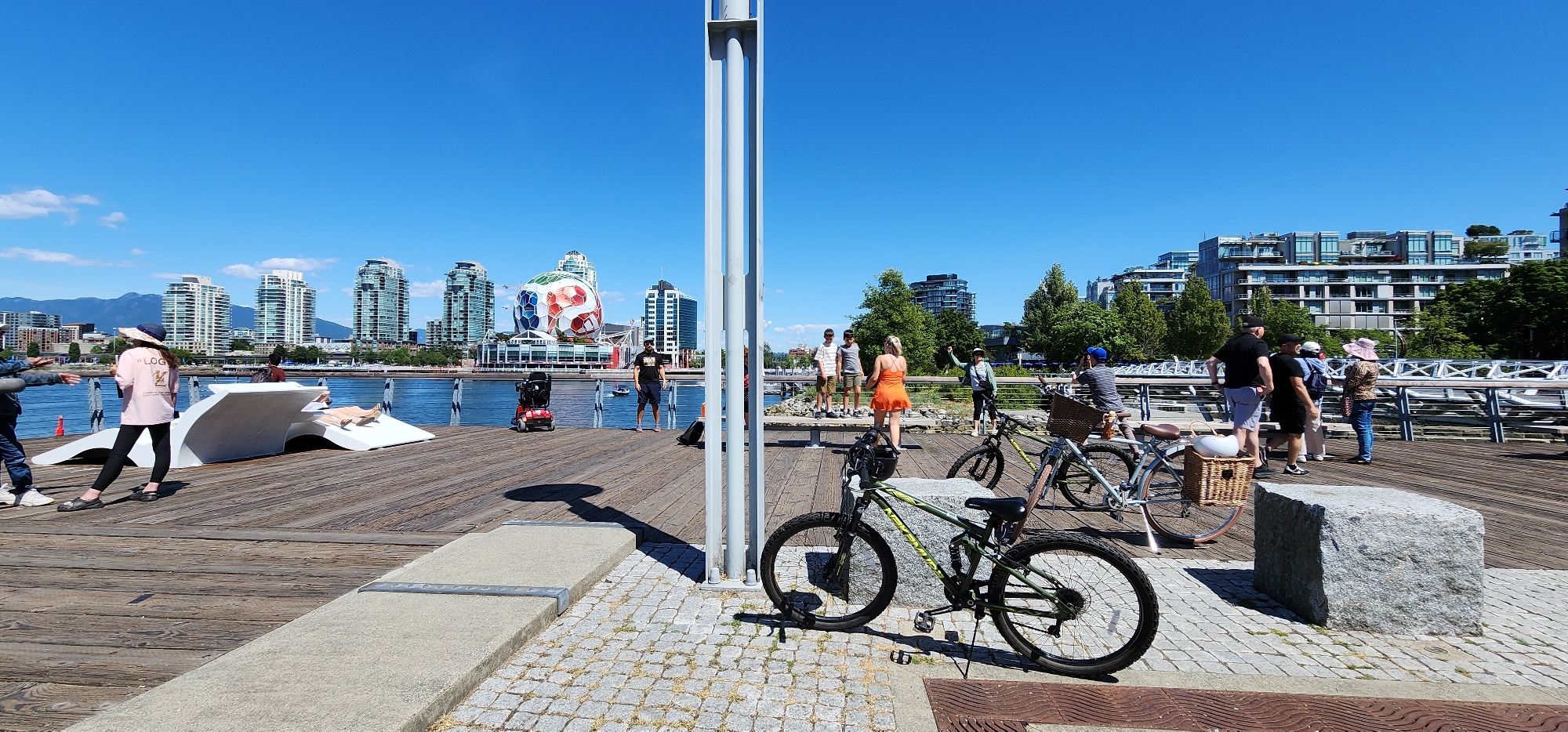 In an outdoor urban waterfront setting, a wide wooden pier is bordered by a body of water on the left and a paved walkway with cobblestone accents on the right. A clear blue sky with sparse clouds dominates the background, with a cityscape of modern high-rise buildings and lush green trees. In the foreground, two bicycles are parked, one green and black, and another lighter colored bicycle with a basket. A large, grey, rectangular stone sits on the cobblestone area.
On the wooden pier, several people are present. A young person wearing a pink t-shirt and dark pants stands near a white sculptural bench. Another person reclines on the bench. A red electric scooter is positioned on the pier. Further down the pier, a group of people are standing and interacting. One person in a bright orange dress is gesturing. Another individual in a blue cap is seated on a bicycle. A group of men and women are engaged in conversation.
In the middle ground, across the water, a large, multicolored, spherical structure resembling a soccer ball is prominently displayed. The cityscape includes various architectural styles of buildings, some with balconies and green roofs. The water appears calm. The lighting suggests it is a sunny day.