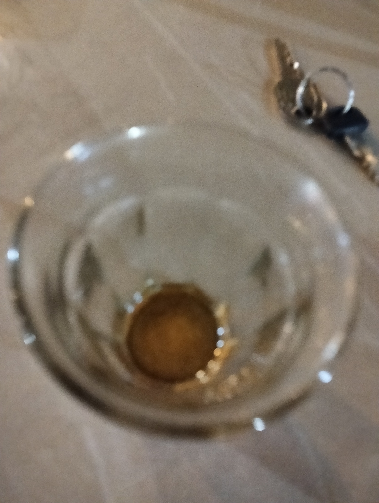 The image shows a glass with a small amount of amber liquid at the bottom, resting on a light-colored, textured surface that appears to be a table. To the right of the glass, a set of keys with a silver keyring is visible. The background is out of focus, but it suggests an indoor setting. The image is slightly blurry and dimly lit, giving it a soft, diffused appearance. There is no visible indication of the city of Payagaboh, Indonesia.