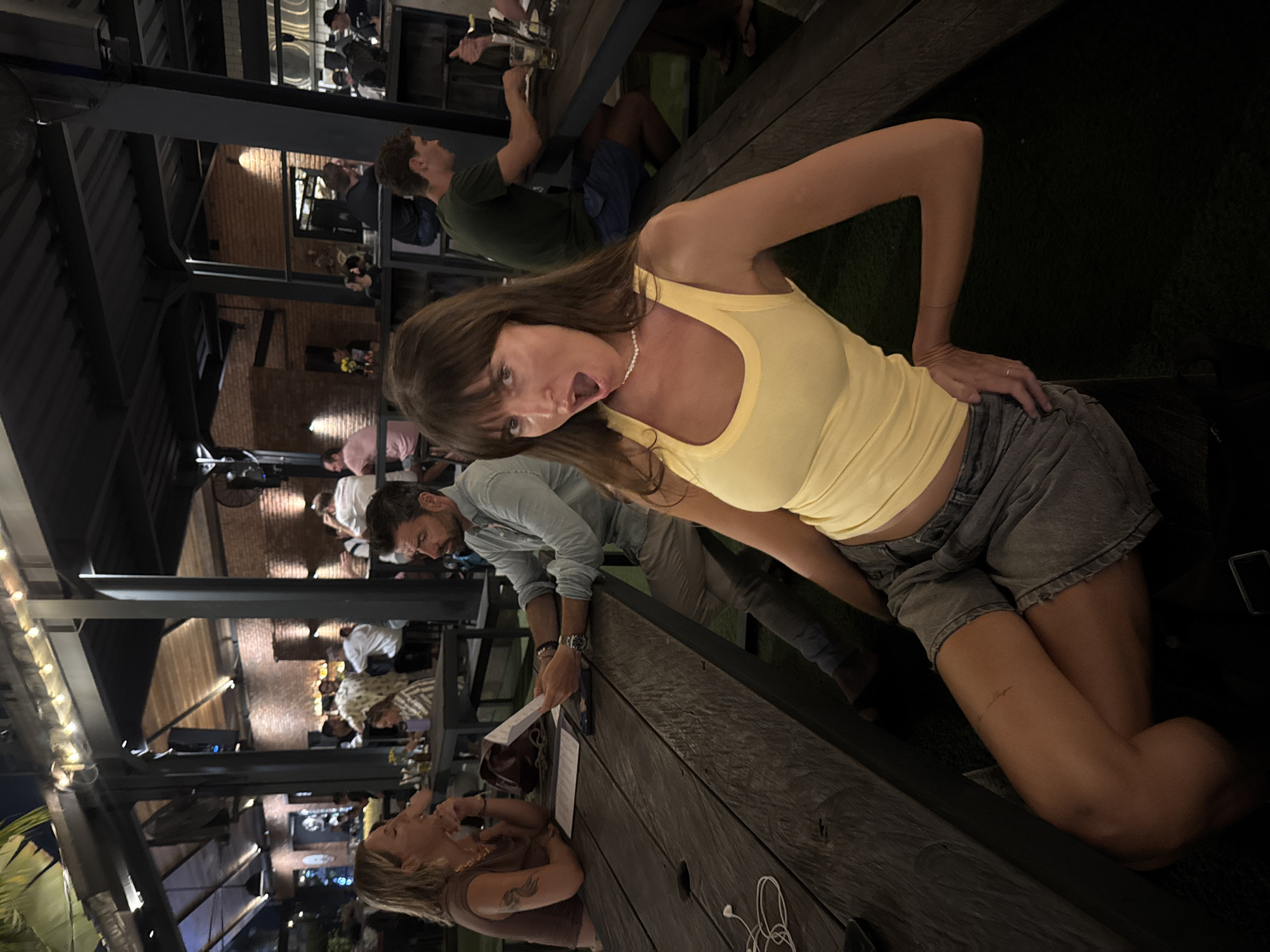 The image depicts an evening scene at a multi-level outdoor or semi-outdoor restaurant or bar, likely in North Kuta, Indonesia. The setting is dimly lit by warm artificial lights, including string lights and spotlights, creating an intimate ambiance.
In the foreground, a woman with long brown hair and bangs, wearing a yellow tank top and light-colored denim shorts, is captured mid-action. She leans towards the camera with a playful or surprised expression, her mouth open, and her right hand resting on her hip. She wears a pearl-like necklace, and a small white smudge is visible on her chin. Her right leg is bent, suggesting she is kneeling or sitting on the edge of a bench.
To her left, at a dark wooden table, a blonde woman in a brown top sits, looking towards a man. A tattoo is visible on the blonde woman's right forearm. The man, wearing a light blue denim shirt and light pants, is leaning over the table, holding white papers, possibly menus or a bill. A watch is visible on his left wrist. A smartphone and tangled white earphones are also on the table.
The background shows multiple other patrons seated at similar dark wooden tables on various levels of the establishment. One man in a dark green t-shirt and blue shorts is visible in the mid-ground. The architecture features dark metal beams and structures supporting the multi-tiered design, with exposed brick walls illuminated by spotlights. Blurred green foliage is visible, reinforcing the outdoor setting. The overall activity suggests people are dining, socializing, and enjoying the lively evening atmosphere. No visible text is legible.