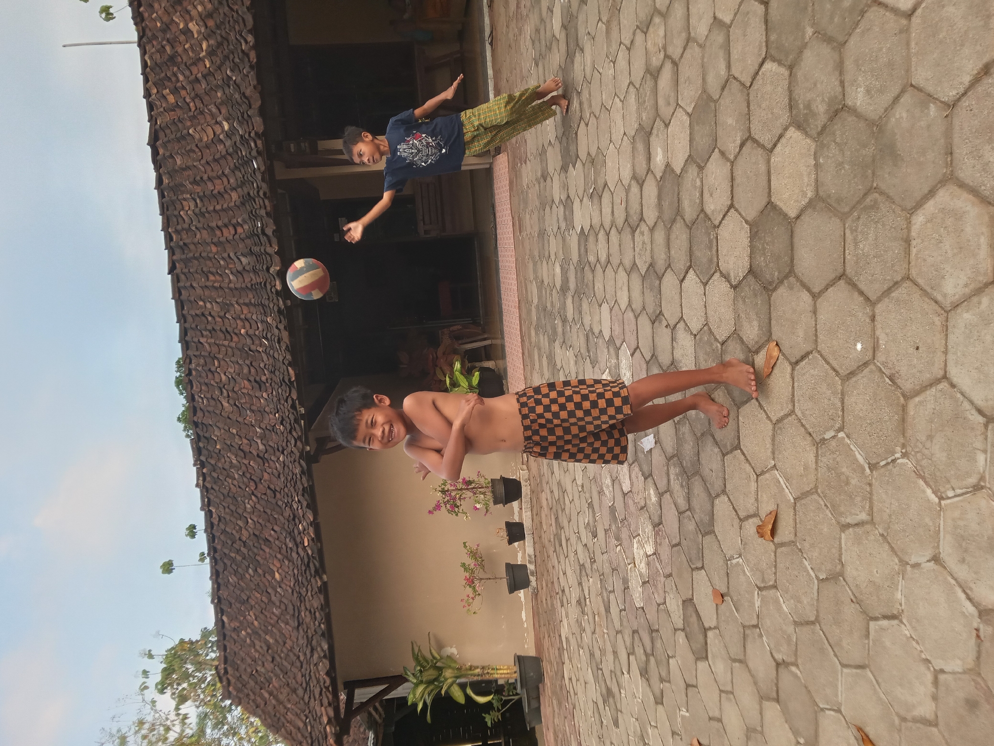 The image depicts two young boys playing volleyball in an outdoor courtyard paved with hexagonal concrete tiles. In the foreground, a shirtless boy with checkered shorts smiles broadly, his arms crossed over his chest. To his right and slightly behind, another boy in a dark blue t-shirt and patterned green and yellow pants is in mid-action, reaching up to hit a volleyball. The background features a building with a tiled roof, potted plants, and some vegetation. The sky is visible above, suggesting it is daytime. The overall scene appears to be a residential area in a tropical climate, possibly Kedunggudel, Indonesia, given the architectural style and lush greenery.