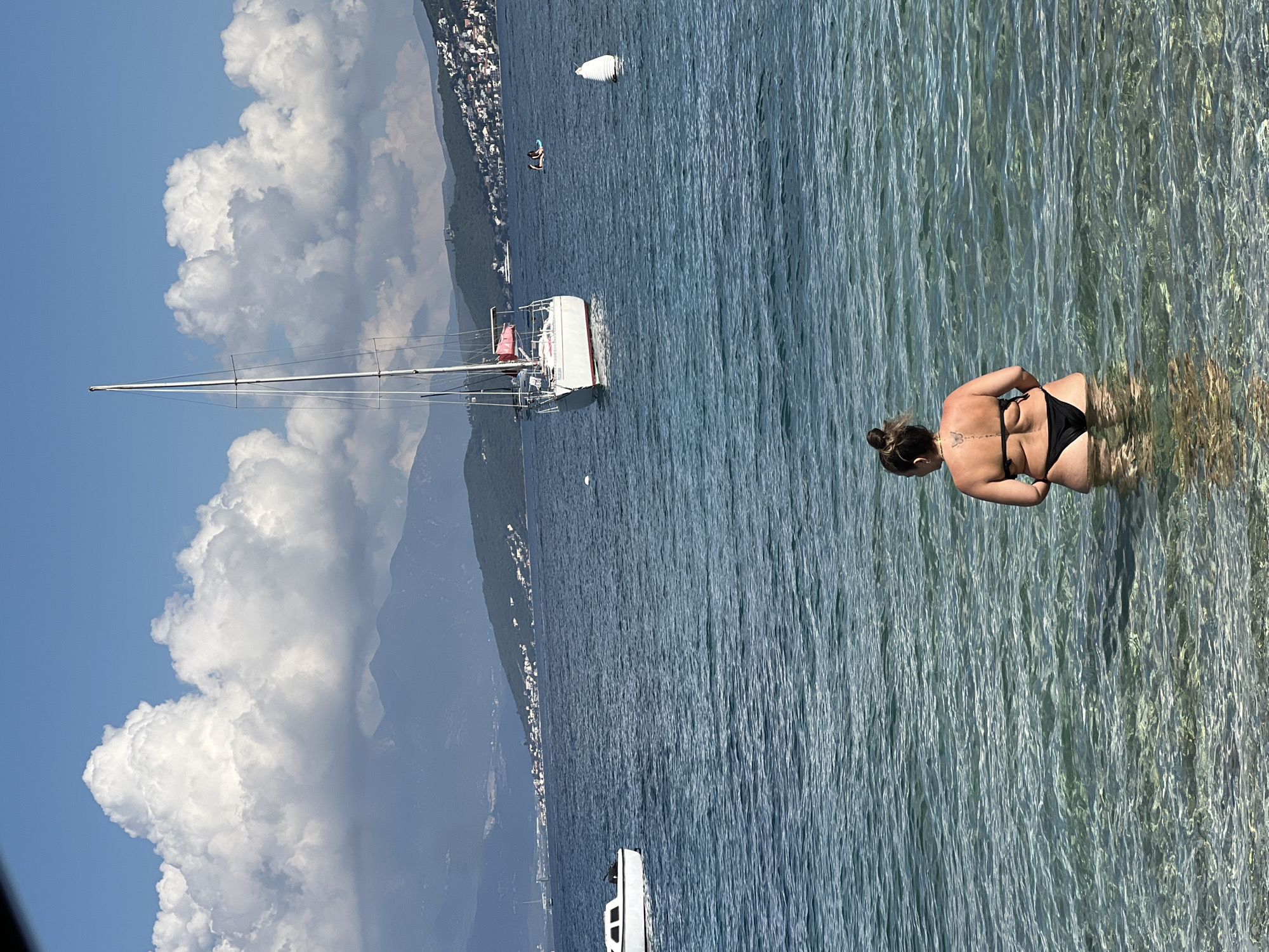 The image presents a serene, sunny daytime scene of a coastal area, likely Đenovići, Montenegro. In the foreground, a woman stands partially submerged in exceptionally clear, shallow water, facing away from the camera. She wears a black two-piece swimsuit, and her dark hair is tied up in a bun, revealing a small butterfly tattoo on her upper back. The clarity of the water allows the sandy bottom to be seen around her.
In the middle ground, a white sailboat with a tall mast and rigging is anchored on the calm, blue waters. Further out, two white buoys are visible, and a smaller white boat can be seen in the far distance near the shoreline. The background is dominated by a majestic, verdant mountainous coastline, densely populated with buildings and houses sprawling along the slopes.
The sky above is a vibrant blue, dotted with large, fluffy white cumulus clouds, indicative of bright and fair weather. The overall impression is one of leisure and natural beauty, characteristic of a pleasant day by the sea. No visible text is present in the image.