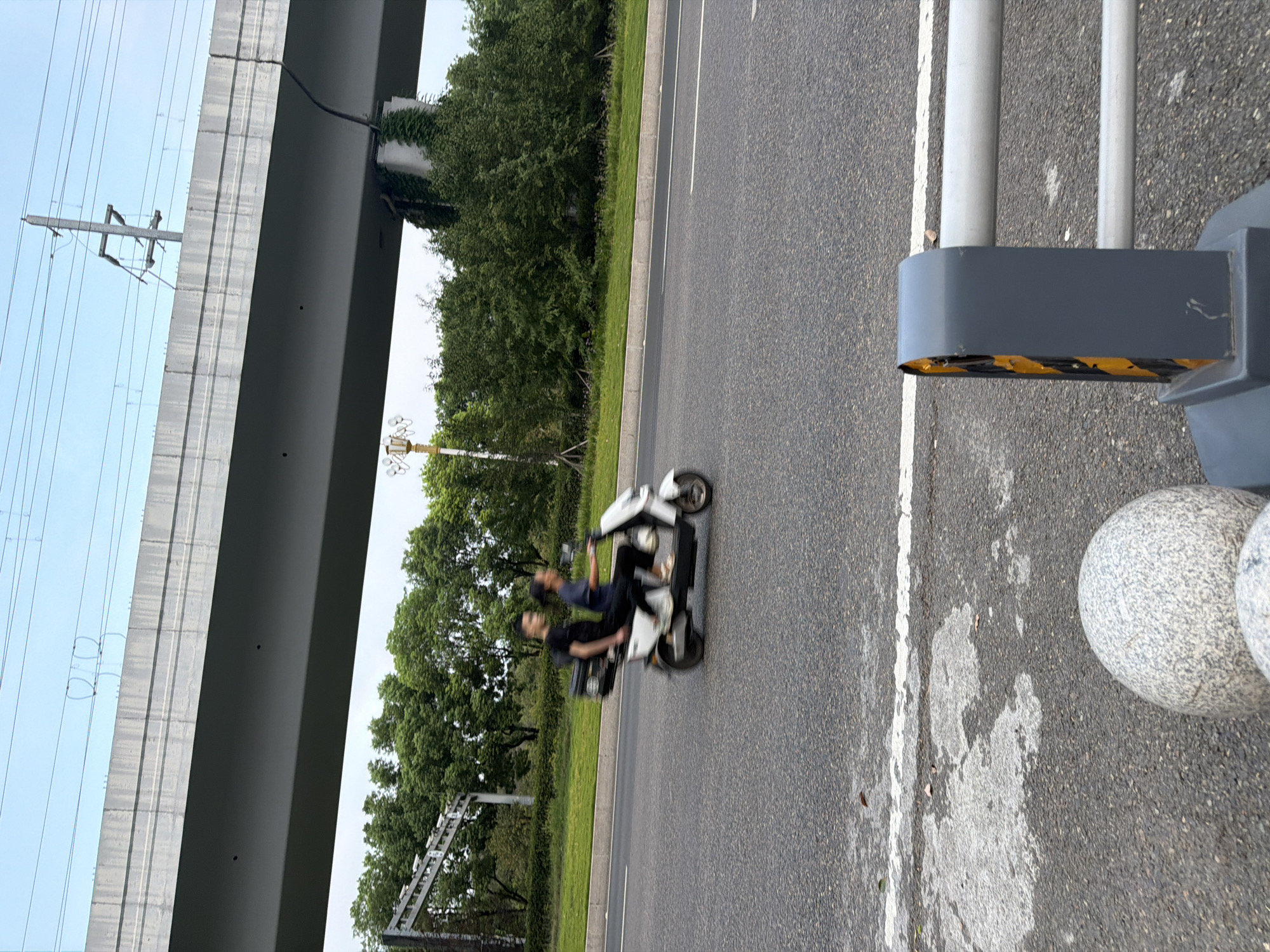 In Jiangjin, China, on an overcast day, a white electric scooter carries two individuals along a paved road beneath an elevated concrete overpass, likely part of a highway or train line. The scooter is in motion, indicated by motion blur. The front rider, wearing a dark t-shirt and pants, steers the vehicle, while the passenger behind, in a dark t-shirt and blue top, is also blurred. The scooter has a rear storage box. The road is bordered by a grassy median and then dense green trees. In the foreground, part of a grey barrier with yellow and black reflective tape and two rounded granite bollards are visible, suggesting a roadside or pedestrian area. Overhead, power lines and poles are strung across the sky, with the overpass casting a shadow.