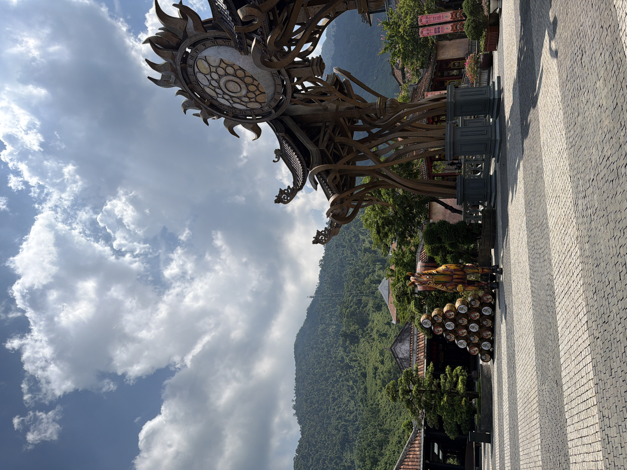 The image shows a view in Đà Nẵng, Vietnam, possibly at Sun World Ba Na Hills. An elaborate metal structure resembling a sun or clock face dominates the upper portion of the frame. Below the metal structure is a tiered, ornamented support. The background reveals a lush, green mountain and sections of buildings and trees. In the lower right, a patterned cobblestone or paved area stretches into the distance, where a colorful statue or sculpture of a horse-like creature stands near a stack of decorated barrels. The sky is partially cloudy with visible blue patches.