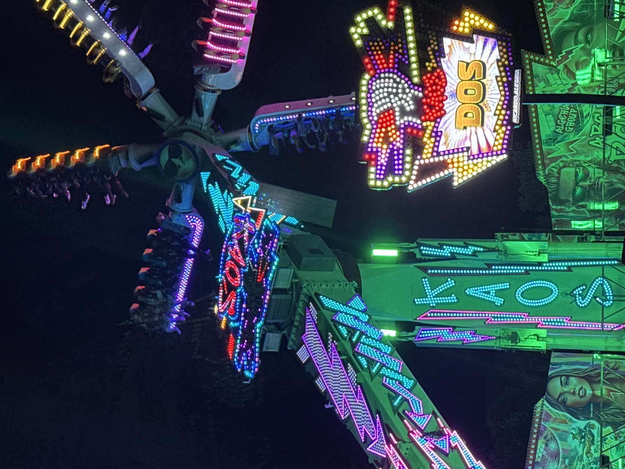 The image depicts a brightly lit amusement park ride at night in Cascais, Portugal. The ride features multiple arms extending from a central hub, each with several pods or seats occupied by people. The structure is adorned with numerous colorful LED lights in pink, purple, blue, yellow, and red, creating a vibrant and dynamic display against the dark night sky.
Prominently displayed on the ride are illuminated signs. One sign, with a dragon-like motif and red and white lights, reads "DOS" in large yellow lettering. Below this, a smaller sign says "RICARDO ALEGRIA". Another sign, running vertically on a green pillar, features the letters "KAOS" in blue and pink lights. In the background, another section of the ride displays lights that spell out "SOAR".
The lower portion of the image shows murals on the side of a structure, featuring stylized portraits of a man with sunglasses and a woman, both rendered in green hues. Text on these murals includes "ALGERIA GROUP". The overall scene suggests a lively nighttime entertainment venue.