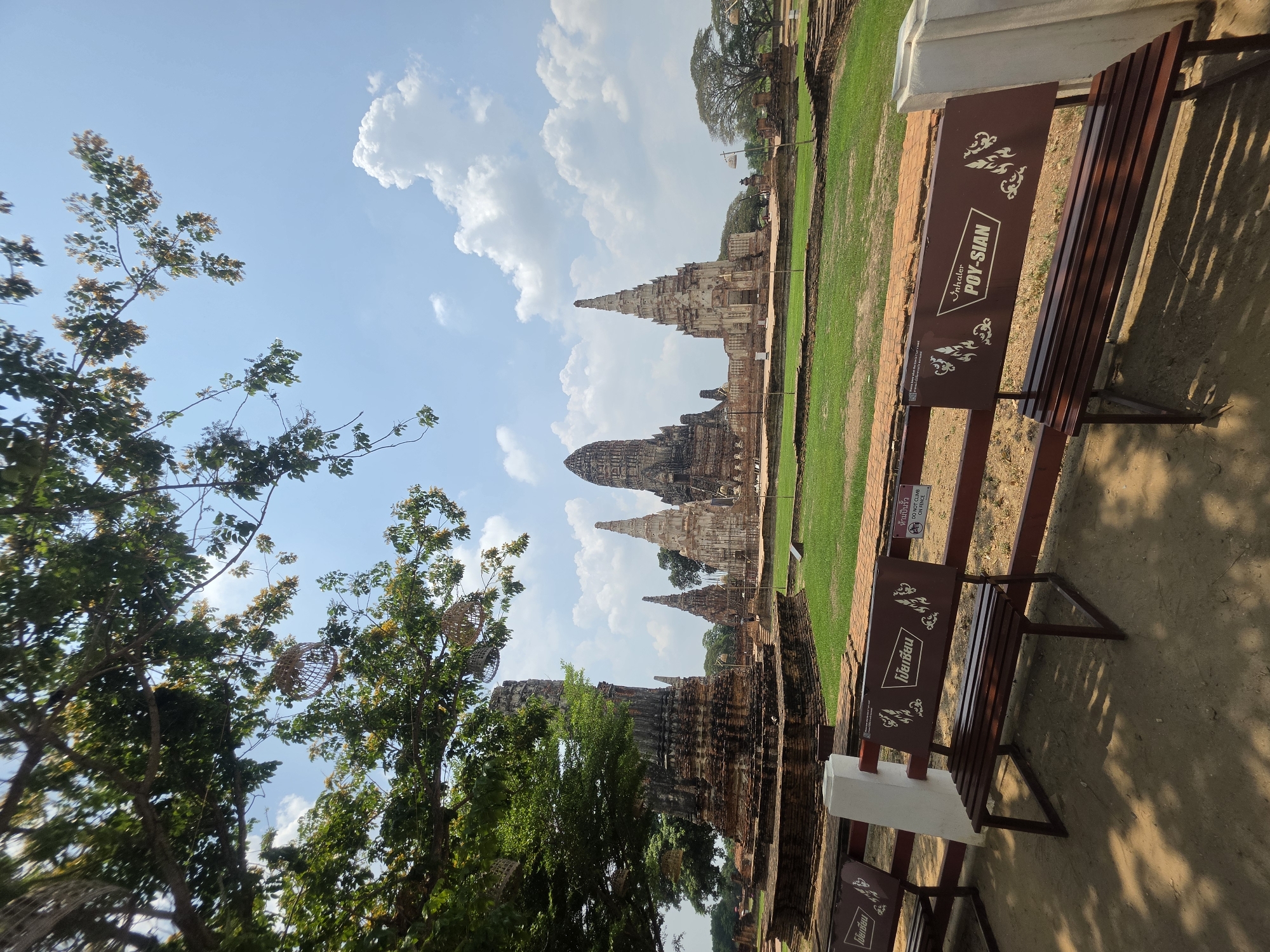 The image shows a view in Ban Pom Subdistrict, Thailand, featuring ancient temple ruins under a partly cloudy sky. Several temple spires rise in the background, surrounded by green lawns. To the right, park benches with signage that reads "POY-SIAN" are present, casting shadows on the ground. Trees with green foliage frame the left side of the image.