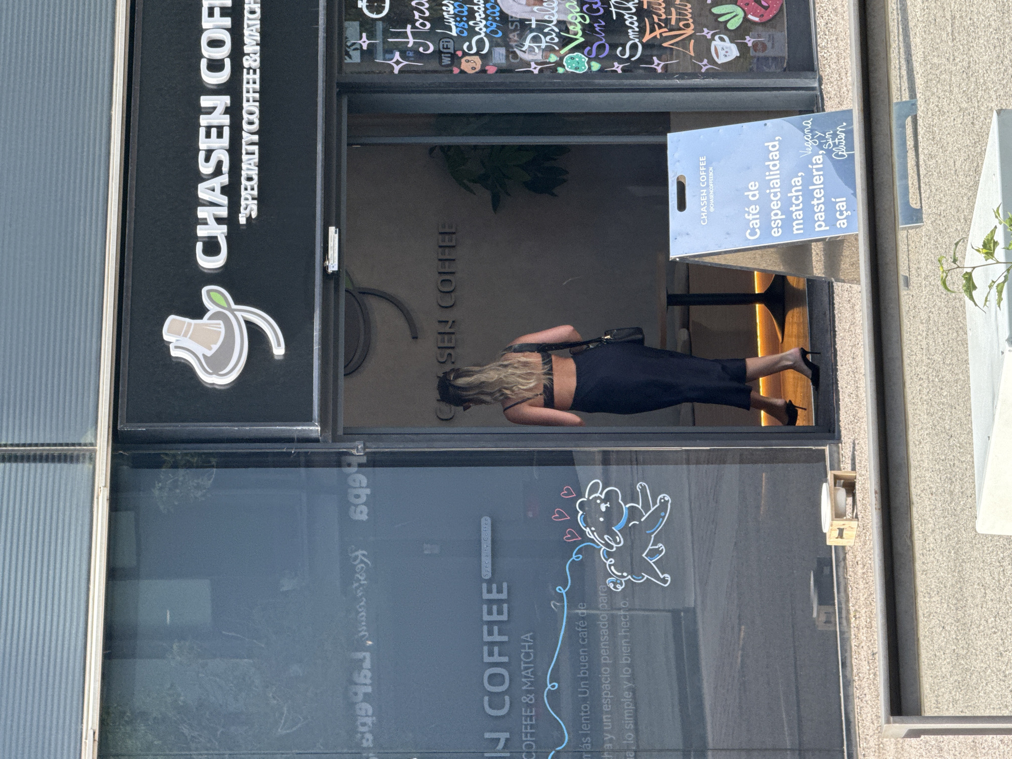 A woman with blonde hair, wearing sunglasses and a black crop top with a matching skirt, walks into a coffee shop. The coffee shop, named CHASEN COFFEE, is located in L'Hospitalet de Llobregat, Spain. It features a large glass facade with reflections of the surrounding area and text. Inside, the shop appears modern and minimalist with dark walls and tables. Outside, a metal A-frame sign displays menu items including "Café de especialidad, matcha, pastelería, acai," and also mentions "Vegana y Sin Gluten." A small wooden platform with a white bowl sits on the pavement in front of the shop.