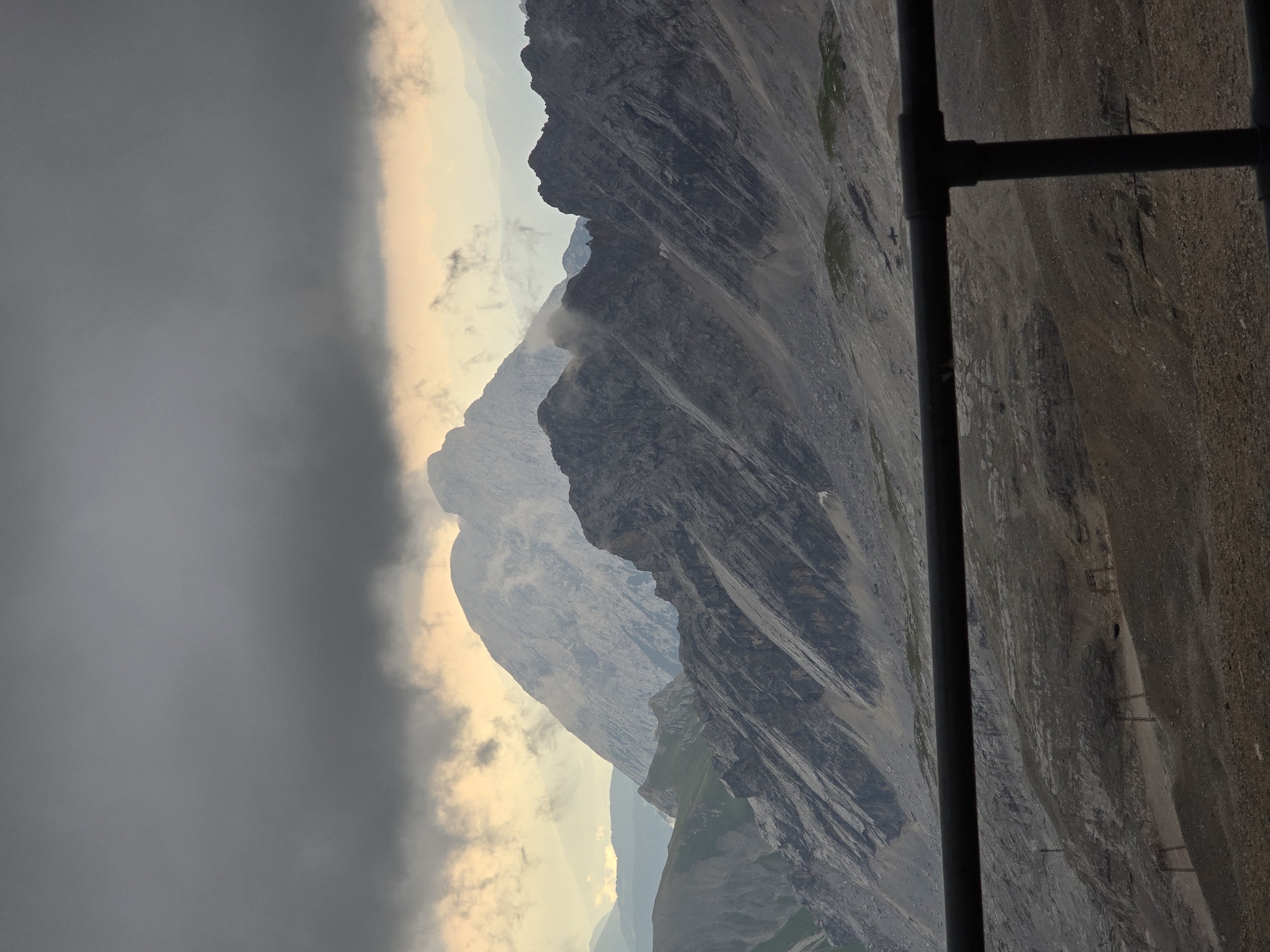 The image depicts a dramatic mountainous landscape under a stormy sky, likely in Garmisch-Partenkirchen, Germany, given the context of its famous Zugspitze peak. In the foreground, a dark railing, possibly from a viewing platform or cable car, partially obstructs the view. Beyond the railing, the terrain is rugged and rocky, with visible paths, some sparse vegetation, and what appear to be remnants of old infrastructure like poles or supports scattered across the slopes. The middle ground is dominated by jagged, dark grey mountain ridges with steep inclines and visible rock striations. Faint wisps of mist or low-lying clouds cling to the upper parts of these mountains. In the background, a massive, imposing mountain peak rises majestically, its lighter grey rocky surface catching the faint glow of sunlight filtering through the heavy, dark grey clouds that dominate the upper portion of the image. The sky is characterized by turbulent, thick cloud cover with a subtle orange and yellow hue near the horizon, suggesting either sunrise or sunset. There are no visible people in the image. The overall impression is one of a formidable and somewhat foreboding natural environment.