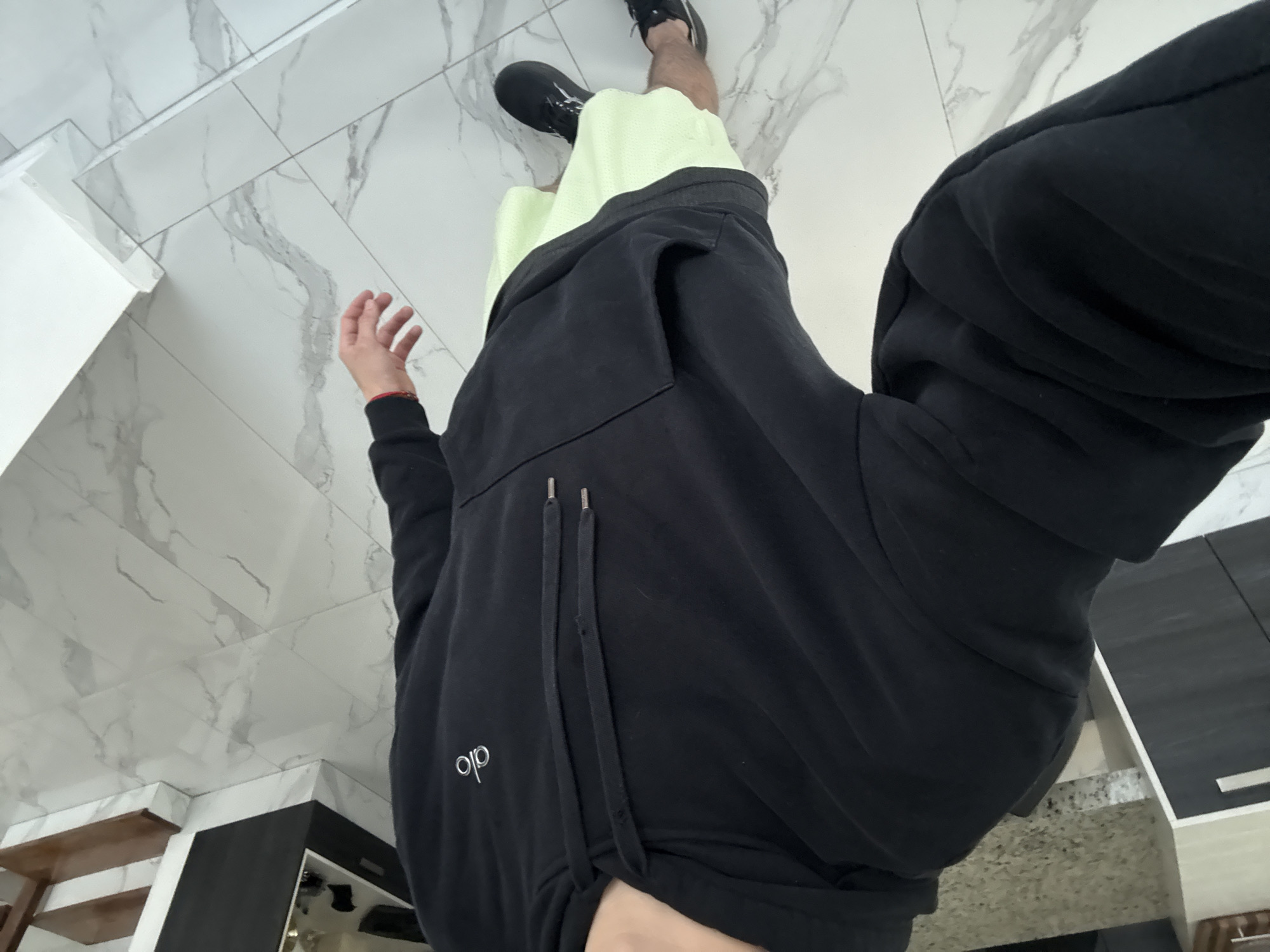 The image displays a high-angle view of a person's lower torso, legs, and feet, situated within a modern indoor environment in San Bernardino Tlaxcalancingo, Mexico. The perspective is from above, looking down.
The subject is dressed in casual activewear. They are wearing black athletic shoes and light green shorts, which are partially covered by a black hoodie or sweatshirt. The black garment features a white "alo" logo on the left side of the chest area and has two prominent drawstrings with silver tips hanging from the collar. A hand is visible on the left side of the frame, wearing a red string bracelet on the wrist.
The setting consists of a clean, tiled floor with a white base and grey marble-like veining. In the background, various indoor elements are visible, including a white wall or partition on the left. To the right, there are modern black and light-colored (possibly wooden or beige) cabinets or shelving units, suggesting a kitchen or contemporary living space. A wooden shelf or counter is also partially visible. The lighting is bright and even, indicating a well-lit interior. The person appears to be standing or subtly moving, possibly posing for a photograph. The visible text is the brand name "alo" on the black garment.