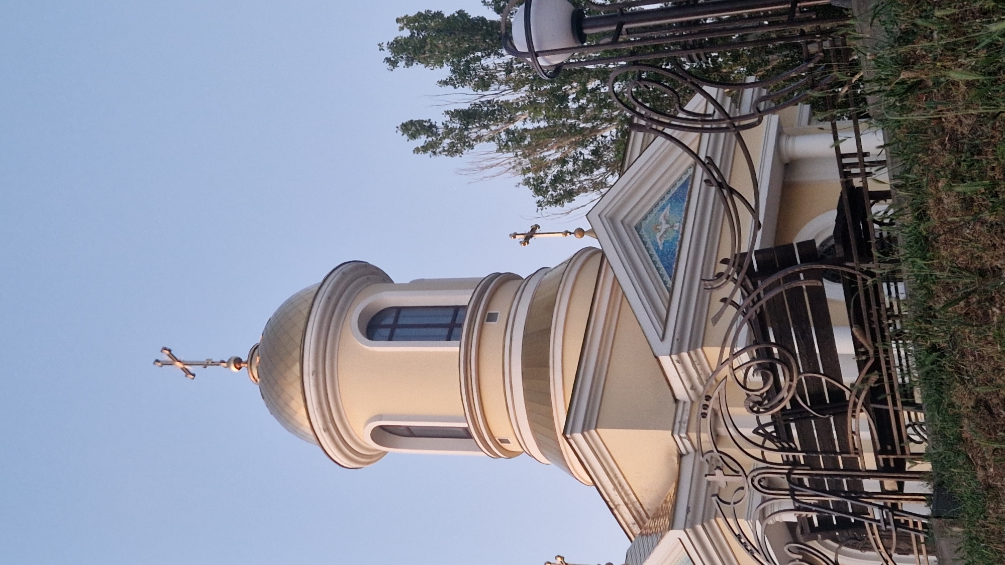 The image shows the exterior of a yellow church with a golden dome topped with a cross. A decorative metal fence and a bench are in the foreground, with grass and foliage. Trees are visible behind the church. The sky is clear and blue.