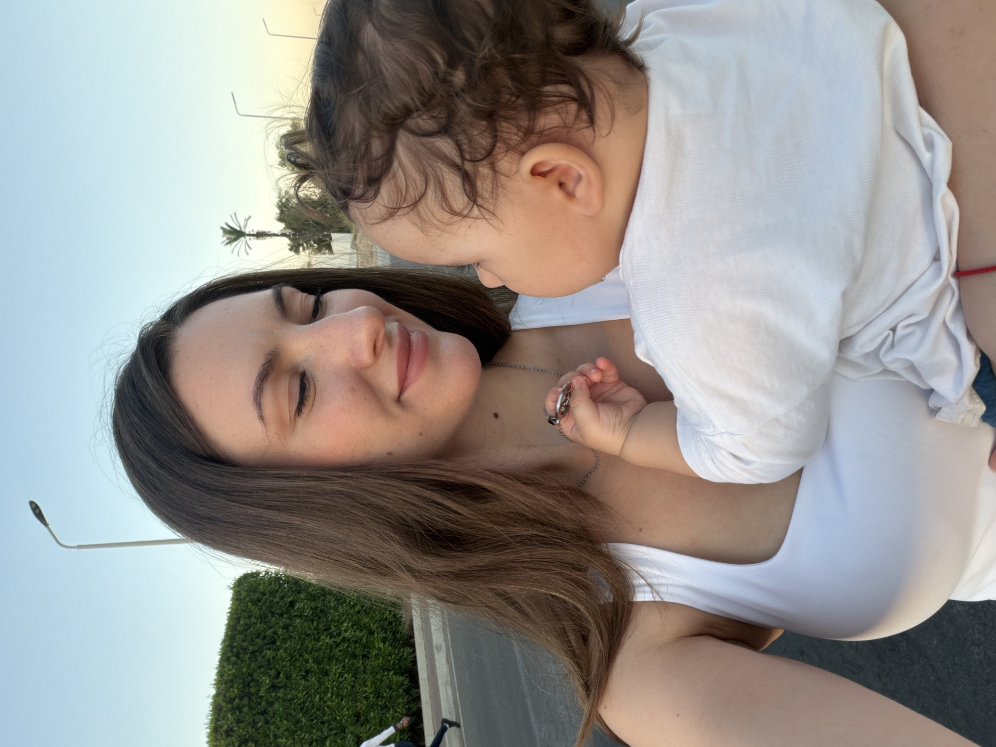 The media file shows a close-up, vertical shot of a woman and a baby outdoors. The woman, with long brown hair, is wearing a white tank top and has her eyes closed, appearing serene or possibly tired. She has a mole on her chest and is wearing a silver necklace. The baby, held by the woman, is wearing a white t-shirt and has dark, curly hair. The baby's head is turned towards the woman, and its hand is reaching towards her necklace.
The background suggests a warm, possibly sunny day, with a bright sky. There are hints of greenery, a palm tree, and a lamppost, consistent with an outdoor setting like a park or a street in a warm climate. The presence of a palm tree and the specified location context of Sharm El Sheikh, Egypt, further solidify this interpretation. There are no visible text elements in the image. The overall mood is one of tender connection between the woman and the baby.