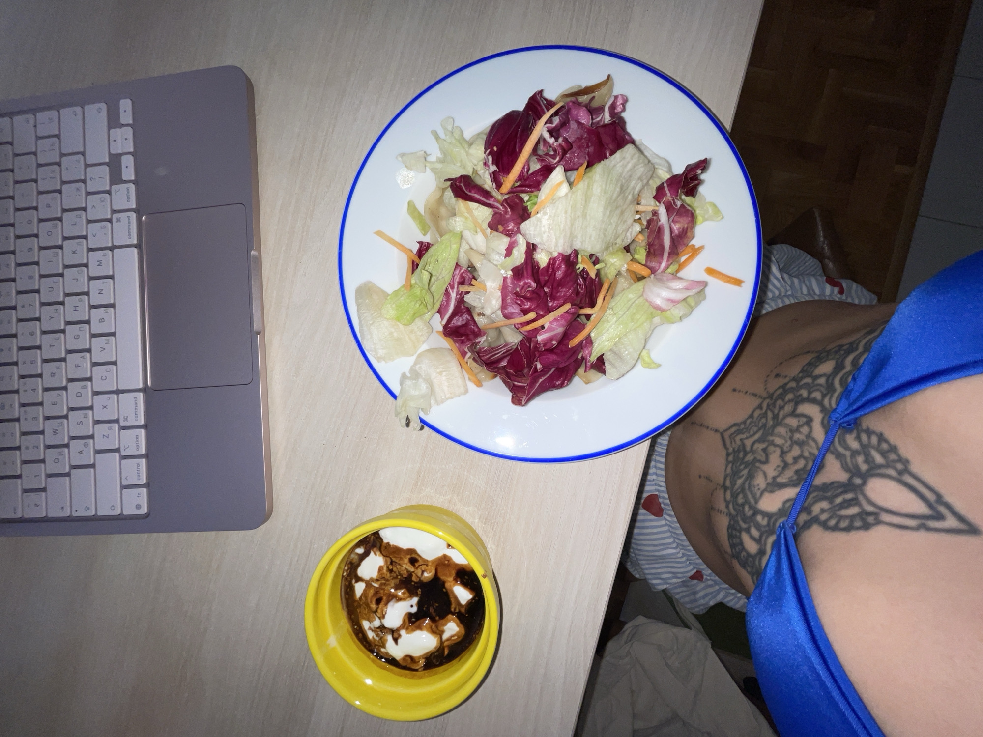 This image captures a casual indoor setting, likely a dining or workspace, in Boreti, Montenegro. The primary subjects include a plate of fresh mixed salad, a small yellow ramekin, and a laptop. A person is partially visible on the right side of the frame, wearing a bright blue, satiny top, with a large, intricate black-ink tattoo visible on their torso or upper back.
The plate holds a vibrant salad composed of light green lettuce, dark red radicchio, and thin strips of orange shredded carrots. Below the salad plate, a bright yellow ramekin contains a dark, glossy liquid with swirls of a white creamy substance, possibly a balsamic glaze or chocolate sauce with cream. To the left, a grey laptop keyboard is visible, featuring both Latin and Cyrillic characters on its keys, indicative of a layout common in regions like Montenegro. Visible text on the keyboard includes "fn", "control", "option", and "command", alongside various letter and number keys.
The activity suggested is a meal or snack break, possibly while working or browsing on the laptop. The lighting appears somewhat dim, suggesting it might be evening or night. The wooden surface beneath the items adds to the domestic or casual atmosphere. No specific emotions are discernible, but the scene conveys a moment of personal time.