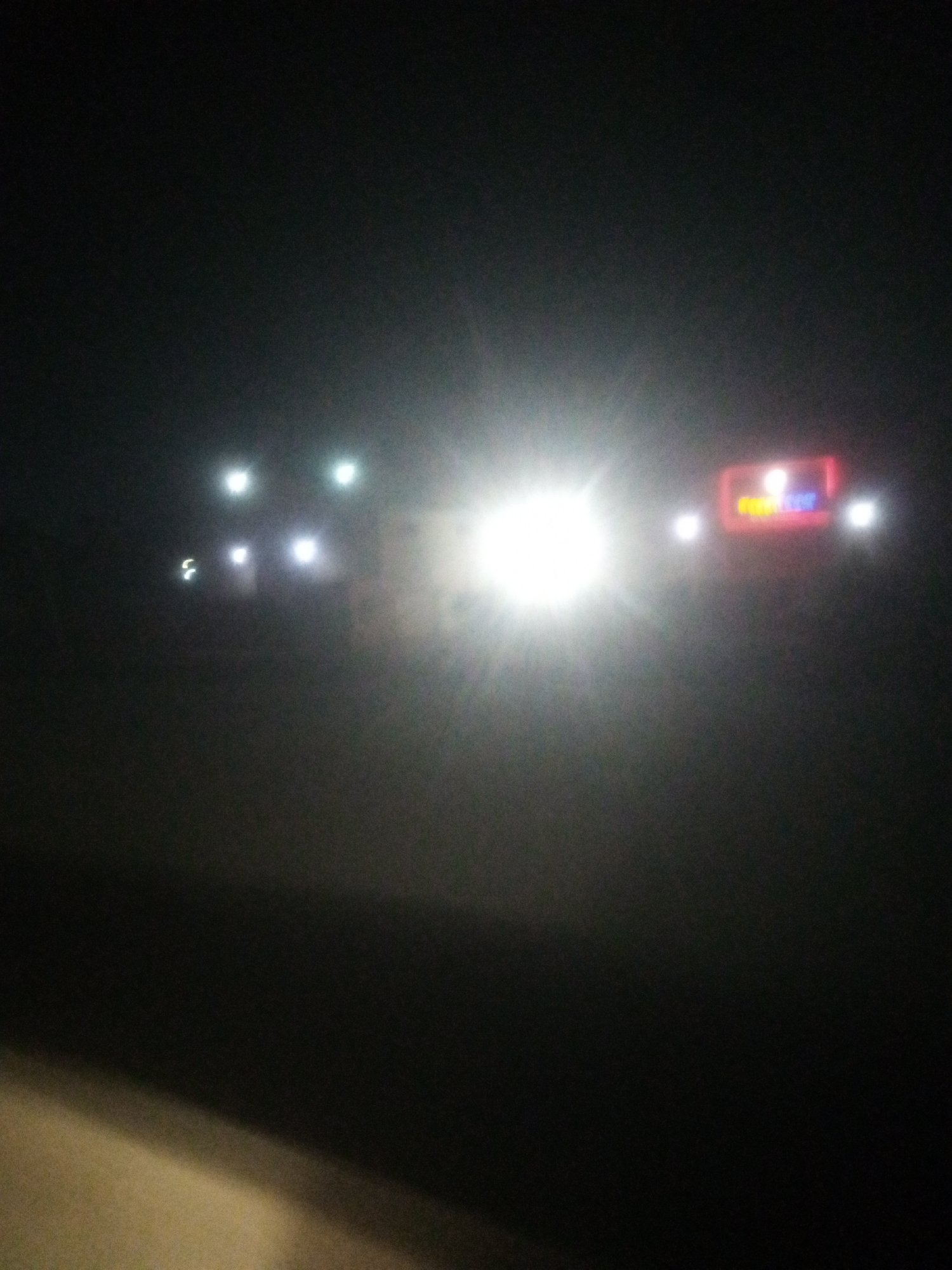 This is a low-light, grainy photograph taken at night in Sabon Gari, Nassarawa, Nigeria. The scene is dominated by a bright, overexposed light source, possibly headlights of an oncoming vehicle, which obscures much of the detail in the foreground and mid-ground. In the background, to the right of the bright light, a neon sign is partially visible, displaying red and blue lettering. The sign appears to be mounted on a building or structure. Several smaller, indistinct lights are scattered in the mid-ground, suggesting other lights from buildings or vehicles. The overall environment appears to be an outdoor urban setting, likely a street, given the presence of vehicle lights and a commercial sign. The ground in the foreground is dimly illuminated, showing a textured surface, possibly asphalt or concrete. The image is characterized by a lack of clear definition and significant glare.