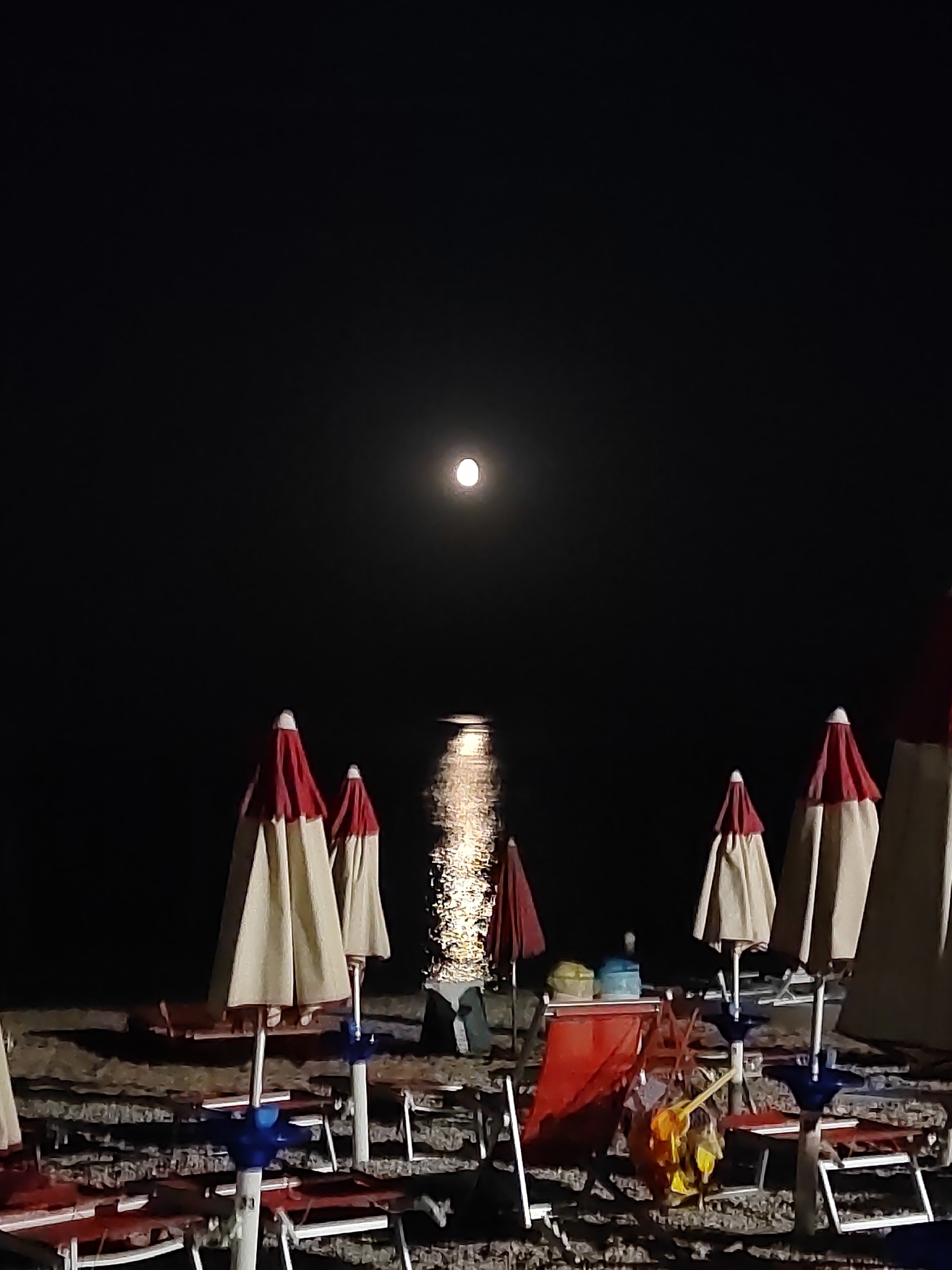 A full moon shines brightly in the dark night sky over the Adriatic Sea, casting a shimmering reflection on the water. On the sandy beach of Porto Recanati, Italy, several beach umbrellas are closed, their red and beige canopies forming cone shapes. Beach chairs are also visible, some with red seats. The scene appears to be deserted, suggesting it is late at night or early morning.