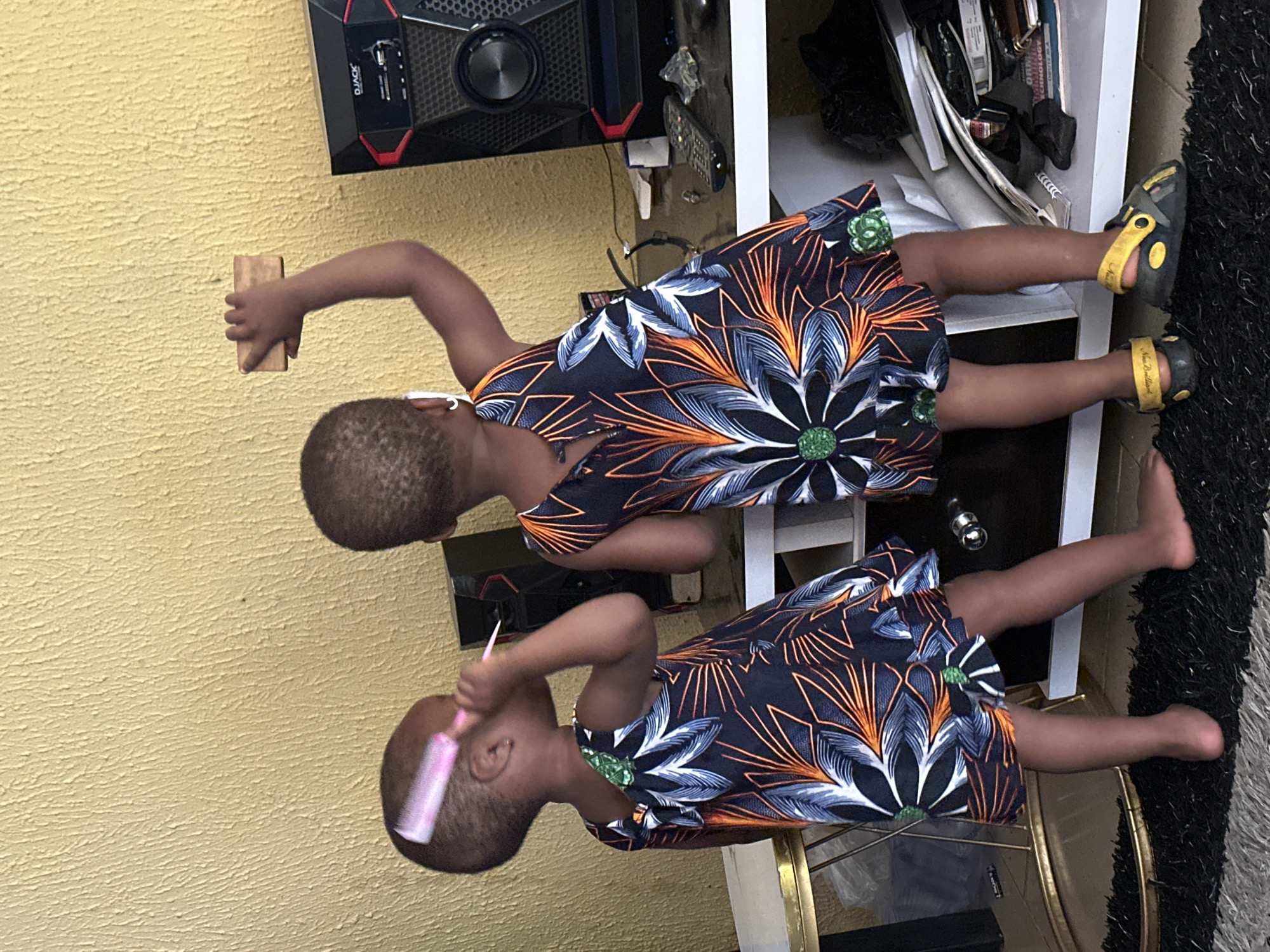 In Narayi, Nigeria, two young girls stand near a yellow textured wall. The girl on the left is wearing a patterned dress and is holding a pink comb to her head. The girl on the right, also in a patterned dress, is reaching up with her right hand to touch a wooden block on the wall. Behind the girls, a white shelf holds various items, including a black speaker with red accents, a remote control, and stacks of paper. The girls are standing on a black shag rug.