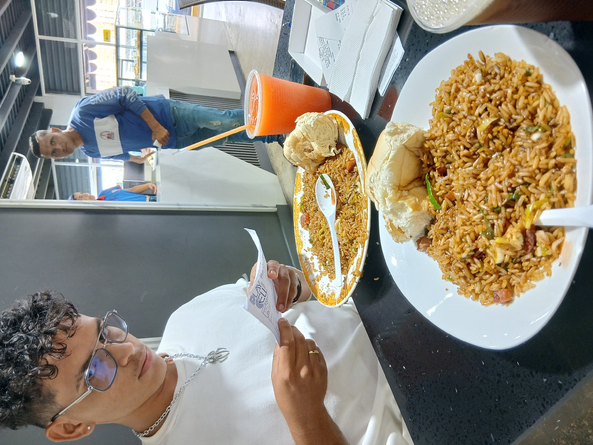 The image captures an indoor scene at a casual eatery, likely during the daytime, in Juan Griego, Venezuela. In the foreground, a young man with curly dark hair and sunglasses is seated at a dark table, intently examining a piece of white paper that bears the text "PLAY CELL." He is wearing a white t-shirt, a silver chain with a spider-like pendant, and a gold ring on his left ring finger.
On the table are two servings of fried rice. One large portion is presented on a white oval plate, alongside a soft white bread roll. A second, slightly smaller portion of fried rice is in a yellow-patterned bowl, also accompanied by a bread roll, with a plastic spoon resting in the dish. Beverages include an orange drink in a clear plastic cup with a straw and a clear glass containing a frothy, light-colored drink. A stack of white paper napkins and various receipts are also visible on the table.
In the mid-ground, a man wearing a blue and white long-sleeved shirt with "SANTA CRUZ" printed on the sleeve and blue jeans is standing, looking towards the right side of the frame, possibly observing a counter or display. A third individual in a blue t-shirt is partially visible further in the background. The setting features bright overhead lighting, large glass windows or partitions, and white interior structures, suggesting a clean and modern environment. The overall activity centers around people dining or waiting to be served.