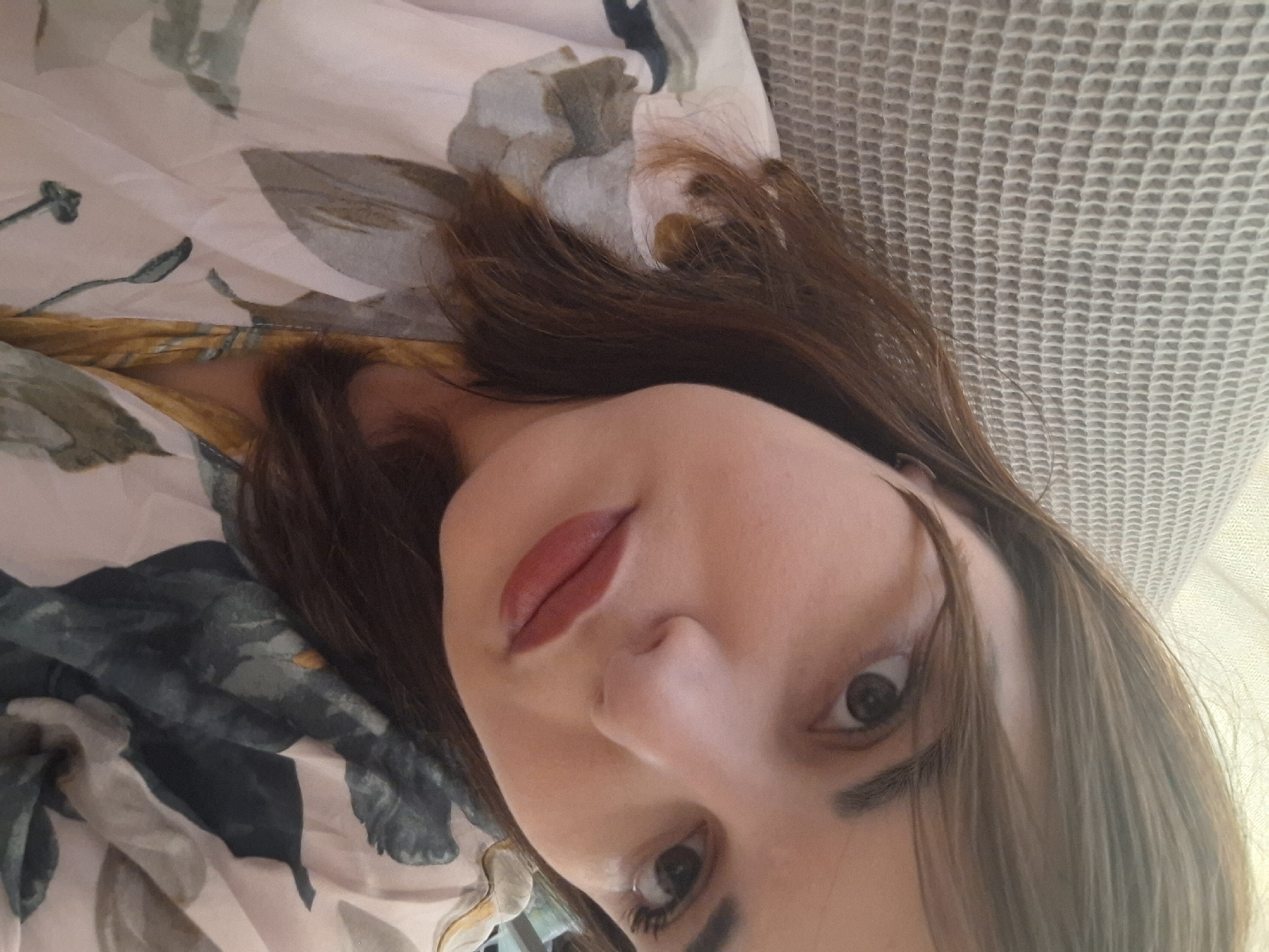 A close-up, tilted portrait of a woman with brown hair and red lipstick. The background consists of a floral patterned fabric and a textured, white surface. The location in the city of Амзя, Russia is unknown.