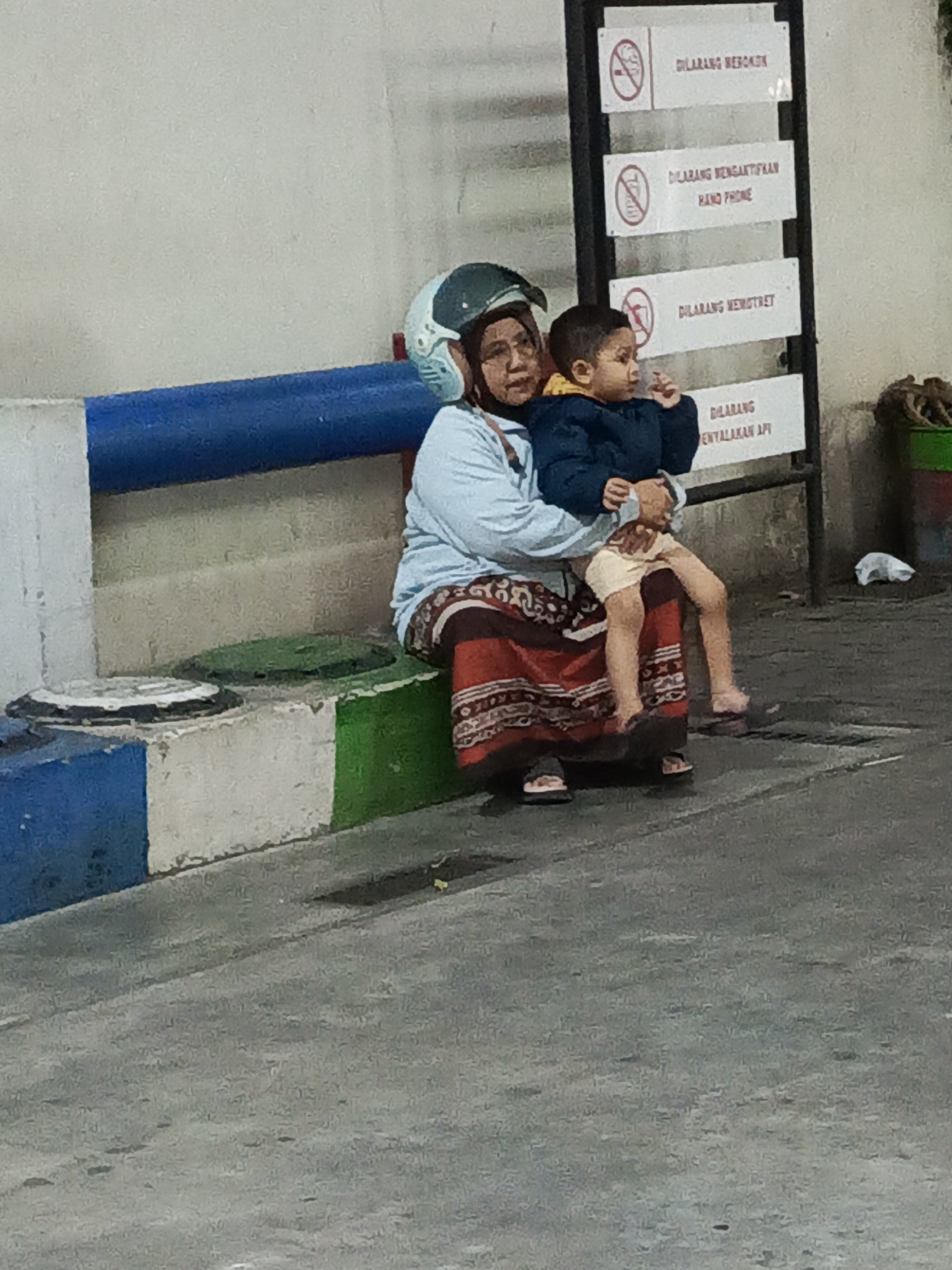 A woman wearing a light blue long-sleeved shirt, a red patterned skirt, and a green and blue helmet is seated on a concrete barrier, holding a young child. The child is wearing a dark blue jacket with a yellow collar and light-colored shorts. They are seated to the woman's right, facing towards the viewer's right. Behind them, a black signpost with multiple white rectangular signs is visible. The signs contain text in what appears to be Indonesian, with red circular prohibition symbols at the top of each. The woman and child are seated outdoors, with a grey paved ground in the foreground and a light-colored wall in the background. To the left of the woman and child, a blue and white striped barrier is visible. The overall scene suggests a public waiting area or a stop, possibly at a gas station or a transportation hub, given the presence of prohibition signs often found in such locations. The environment appears to be urban, with a neutral color palette.