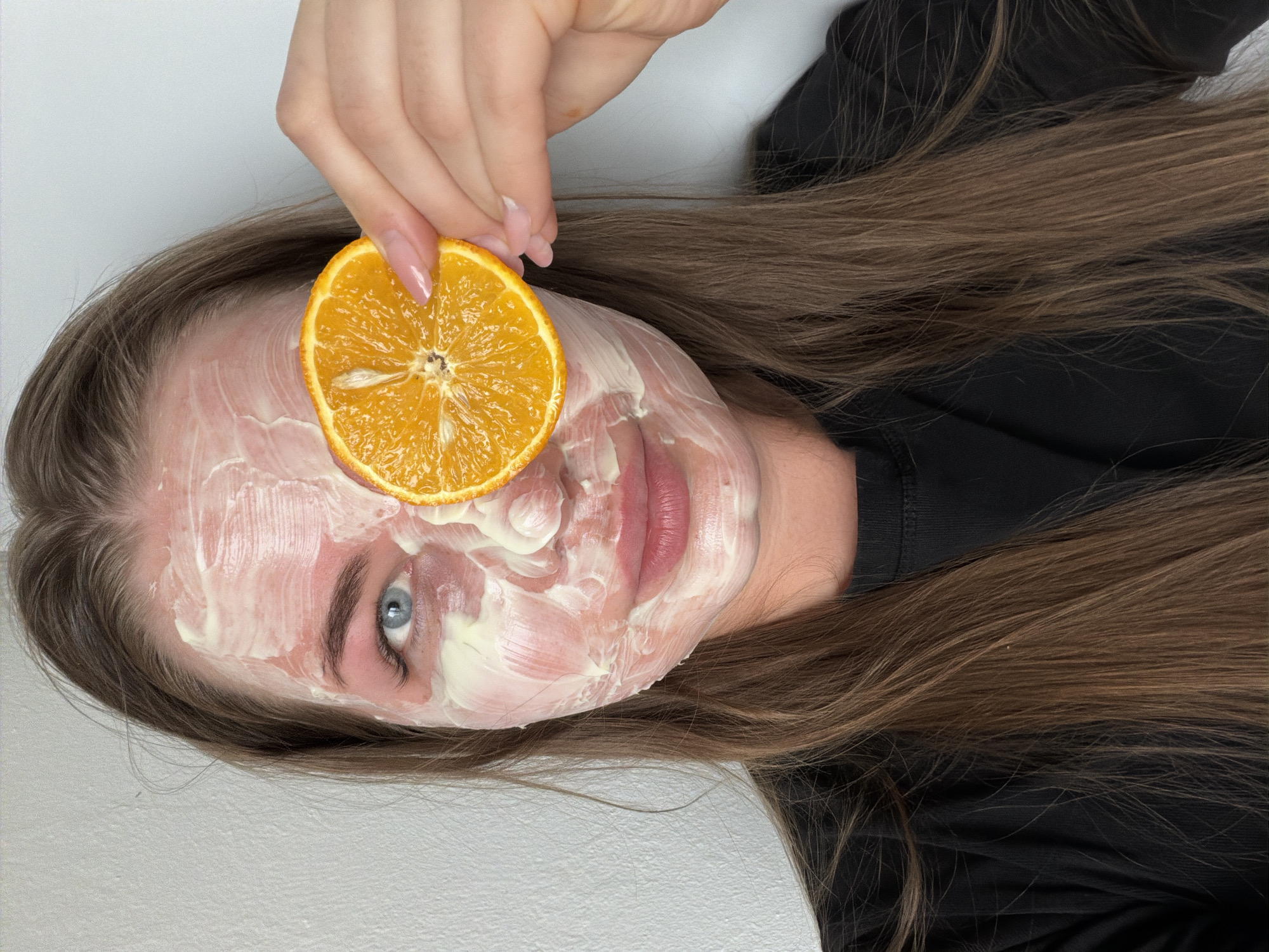 A fair-skinned woman with light brown hair and blue eyes is lying on a white surface, possibly a bed or a table, wearing a black shirt. She has a thick, white facial mask applied. She is holding a slice of orange over her face with her right hand.
The setting appears to be an indoor environment. There is no specific information to confirm the location as Chaikovsky, Russia.
