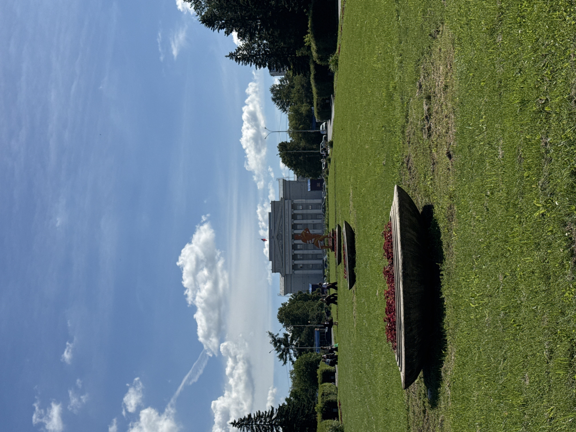 This image depicts a wide view of a grassy park area with a large, classical-style building in the background. The building features a colonnade and a flag on its roof. In the foreground, there are two large, shallow planters, one of which contains red flowers. Several people are visible walking in the middle ground, and trees line the perimeter of the park. The sky is blue with scattered white clouds. This scene is identifiable as being in Yekaterinburg, Russia, specifically at the Yekaterinburg City Administration building, located at 248 Malysheva Street.
