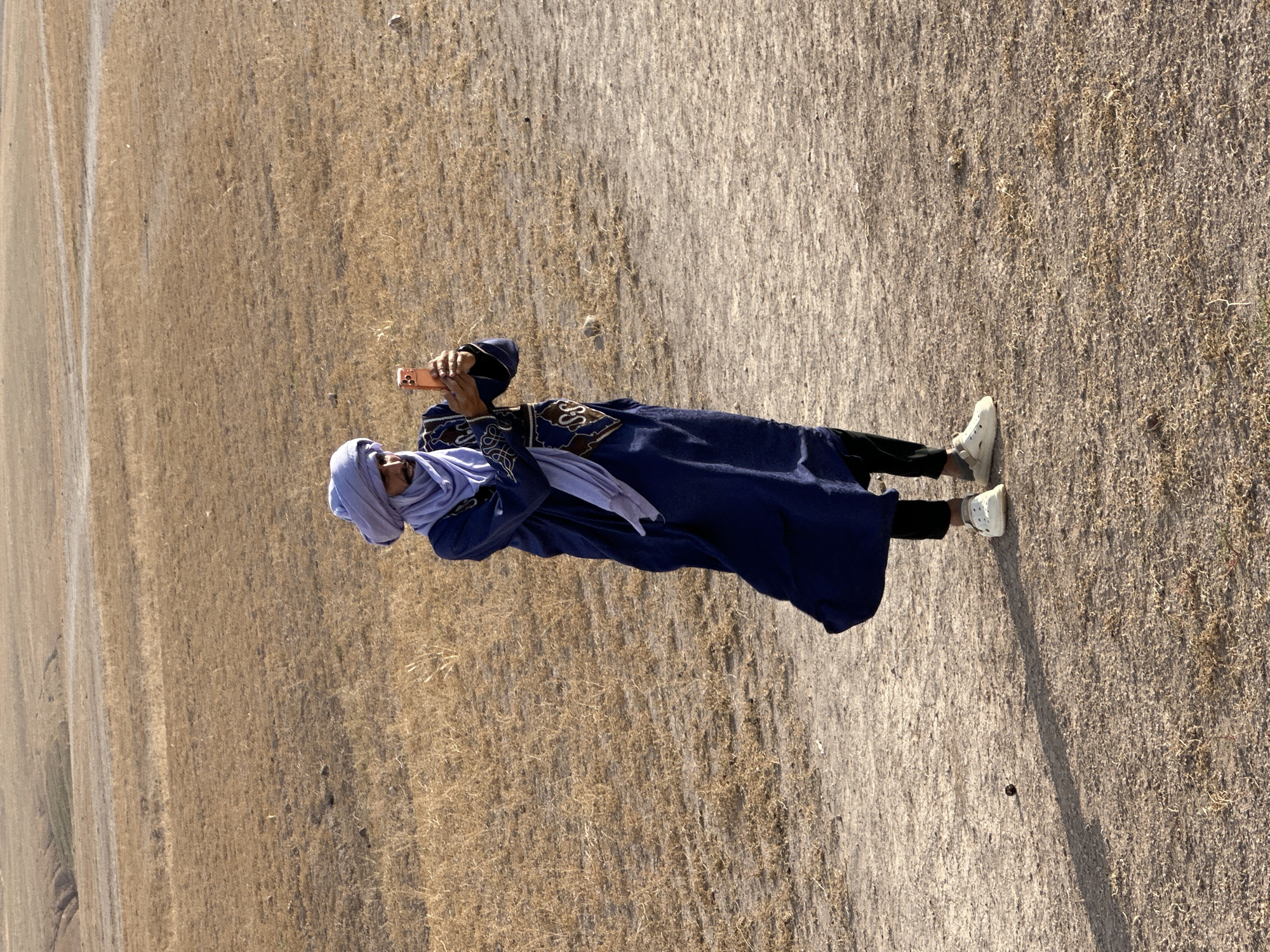 A person in traditional Moroccan attire stands in a field, holding a mobile phone and taking a picture. The person is wearing a blue robe with gold embellishments, black pants, white shoes, and a light blue head covering. The field is dry and grassy, with a dirt path visible. The location is in Sidi Badhaj, Morocco.