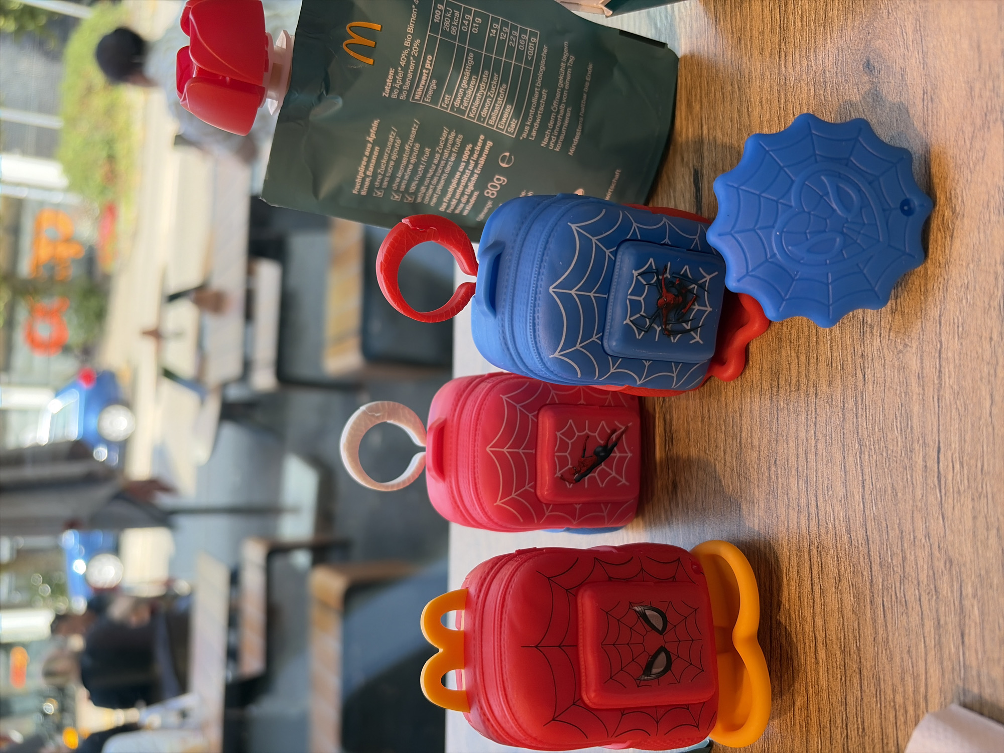 In the city of Langenthal, Switzerland, this image captures a close-up view of three McDonald's Happy Meal toys, designed as mini backpacks, arranged on a wooden table. The toys feature a Spider-Man theme. The leftmost toy is red, the middle toy is pink, and the rightmost toy is blue, all adorned with a white spiderweb pattern and the iconic Spider-Man logo. In the background, out of focus, are tables and chairs of an outdoor dining area, suggesting a McDonald's restaurant setting. A green pouch, likely containing fruit puree, is also visible to the right of the toys, displaying McDonald's branding and nutritional information in German. People are faintly visible in the blurred background, engaged in outdoor dining activities.