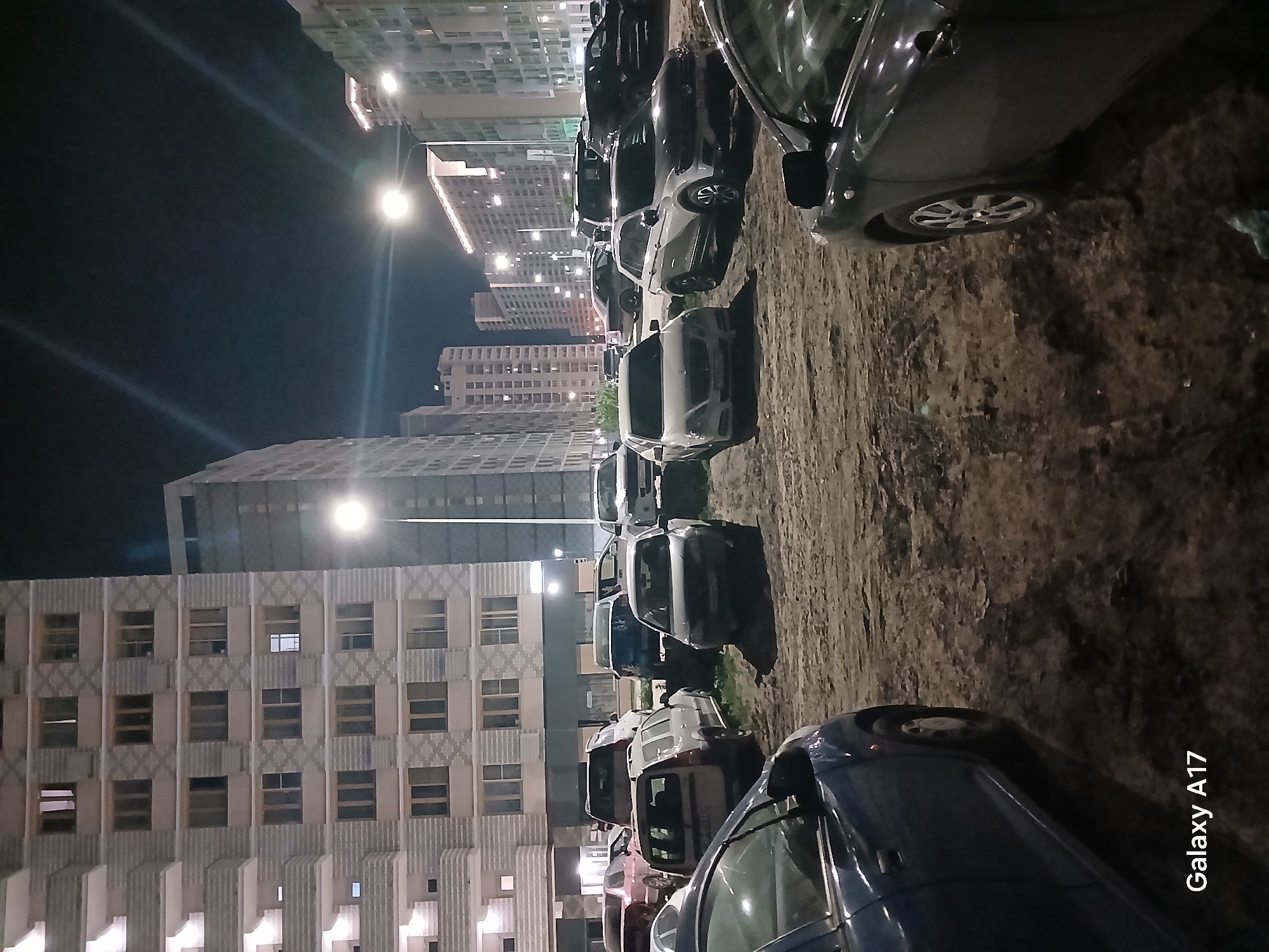 The image depicts a nighttime scene in an urban environment, likely a residential area in Yekaterinburg, Russia. In the foreground, a row of parked cars stretches into the distance. The vehicles are a mix of colors and models. The ground appears to be dirt or gravel.
In the background, tall apartment buildings dominate the skyline. Streetlights illuminate the scene, casting light on the buildings and cars. The buildings exhibit a repetitive pattern of windows and architectural details. The overall atmosphere is quiet and still.