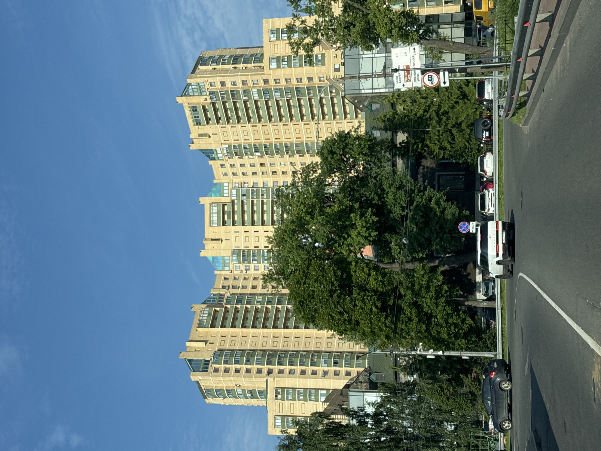 A wide-angle, exterior, eye-level, medium shot captures a modern, multi-story yellow apartment building with green accents and balconies, dominating the background under a clear blue sky with sparse clouds. In the mid-ground, a large, mature green tree obscures a portion of the building. The foreground shows a paved road with a white dividing line, a metal guardrail, and several vehicles including a white SUV and a dark sedan. Several traffic signs are visible on the right side of the road. The overall scene appears to be in a developed urban environment.