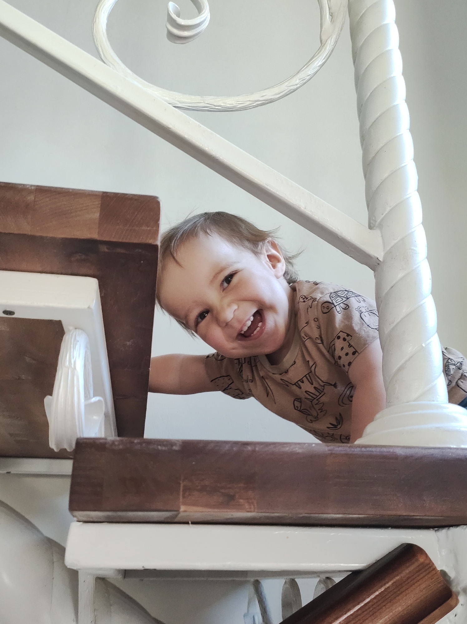 A young child is positioned on stairs, appearing to peek from behind a landing. The child is smiling broadly, with an angled head and visible teeth. The child's clothing is a beige t-shirt patterned with cartoon-style drawings. The stairs are constructed with dark-stained wooden treads and white railings, including a decorative, rope-like column. The background is a plain, light-colored wall. The location is Unknown, Unknown.