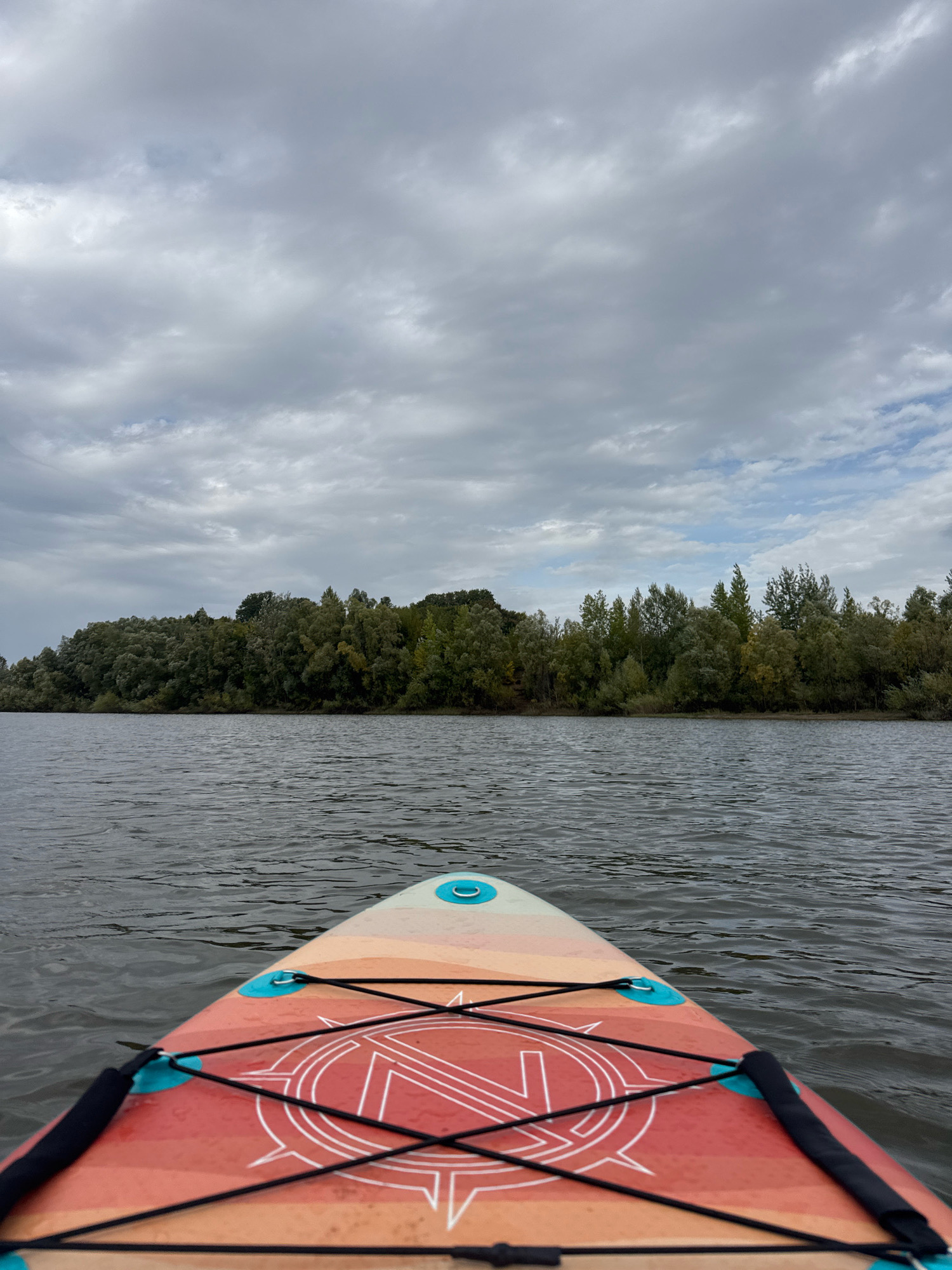 A first-person perspective view from a paddleboard shows a calm body of water. The paddleboard's nose, adorned with orange and white stripes and a compass-like design, occupies the foreground. Black bungee cords crisscross the paddleboard for securing gear. Beyond the water, a dense line of green trees forms a natural shoreline, with some trees showing hints of autumnal yellow. The sky above is overcast with dramatic grey clouds. This scene is likely from the Ufa area of Russia, given the vegetation and typical weather patterns. No people are visible.
