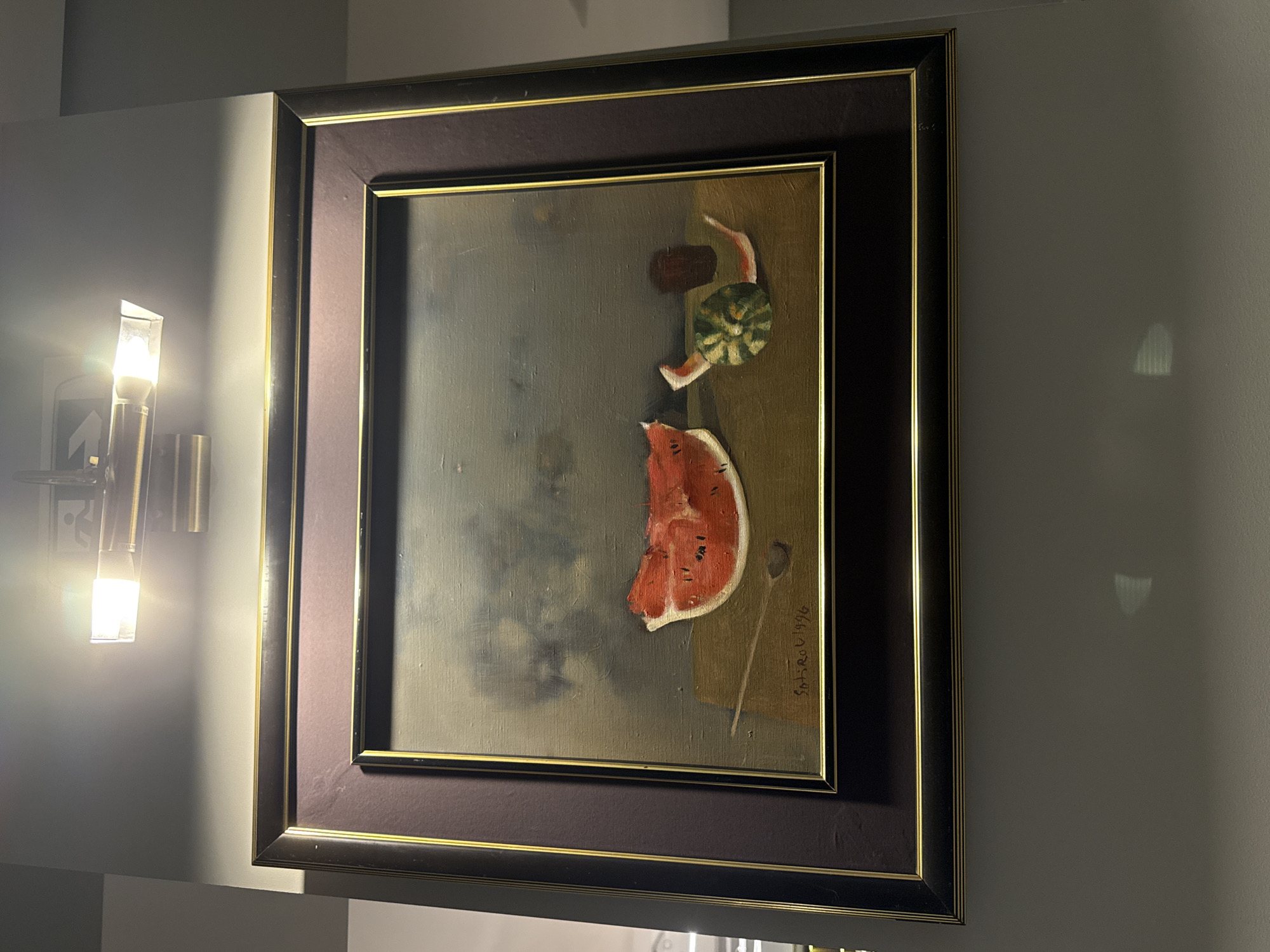 The image shows a framed painting of a still life featuring watermelon. The painting is illuminated by a bright light fixture above it, casting shadows on the wall and the artwork itself. The painting depicts a slice of watermelon and a smaller, halved watermelon with rind, placed on a wooden surface. A signature and date, "SATIR OU 1996," are visible in the lower left corner of the painting. The painting is hanging on a light gray wall. The overall setting appears to be indoors, possibly in a home or gallery. The city of Budva, Montenegro is not visible or implied in the image.