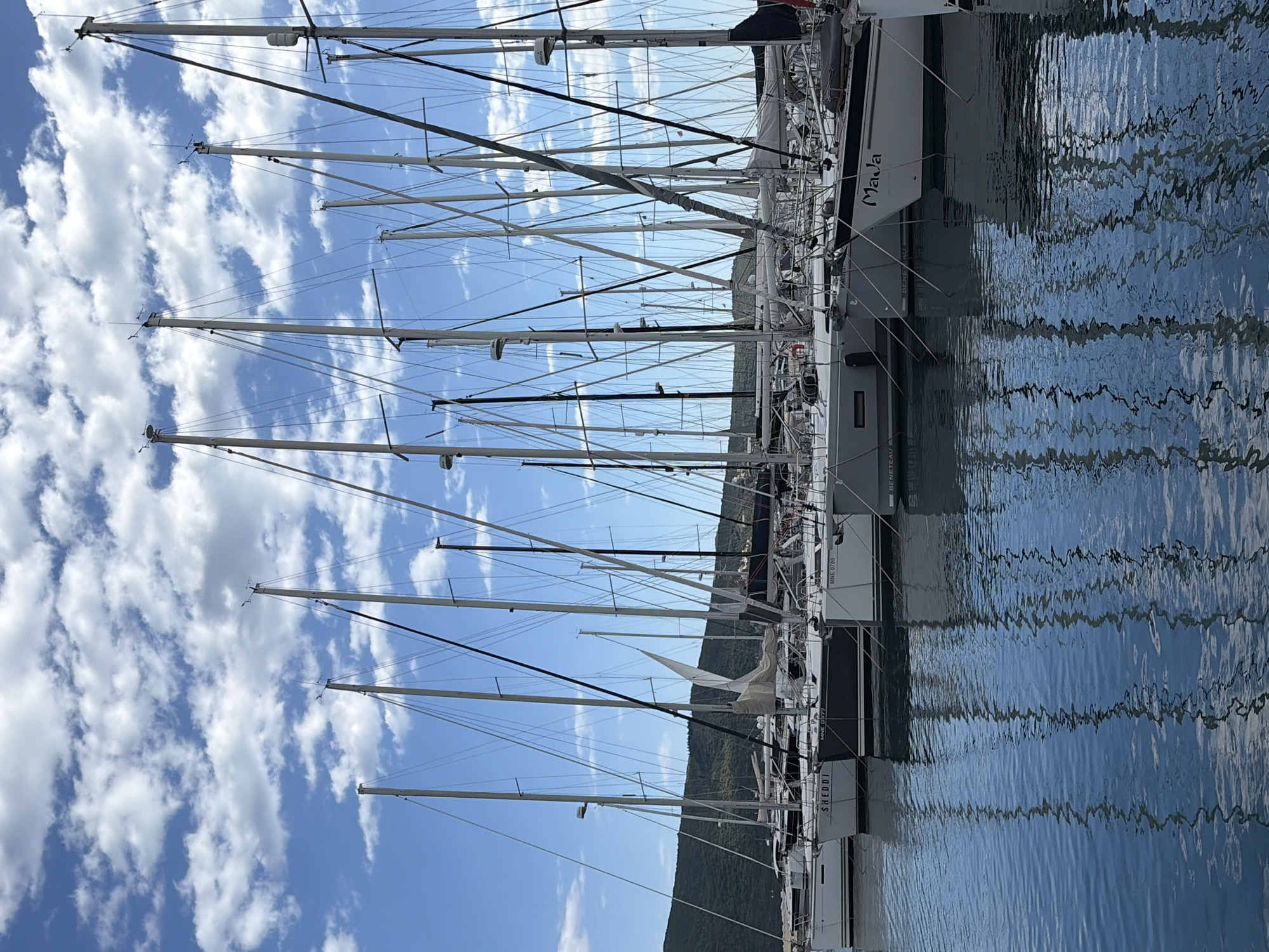 The image captures a marina scene in Meljine, Montenegro. Numerous sailboats are docked, their masts reaching high against a backdrop of a bright blue sky scattered with white, puffy clouds. The boats are aligned side-by-side, reflecting in the calm water of the marina. A verdant, low-lying mountain range forms a distant backdrop. The reflections on the water create a rippled effect, distorting the image of the boats and the sky. The name "Mala" is visible on one of the boats. The overall scene suggests a peaceful, sunny day at a coastal marina.