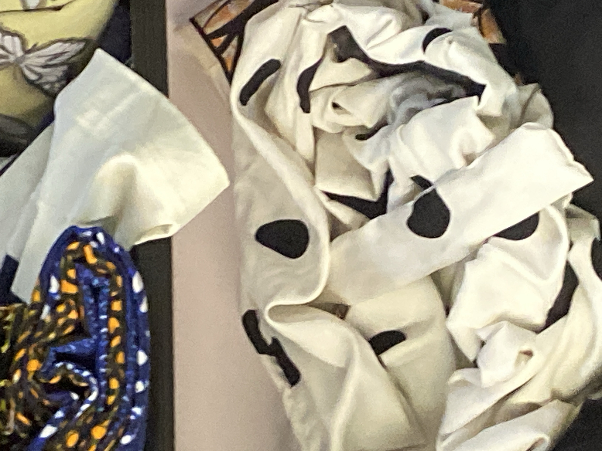 This is a close-up, slightly out-of-focus photograph of folded fabric. The fabric appears to be of two types. In the upper left corner, a tightly rolled piece of fabric displays a pattern of small orange, yellow, and white dots on a dark blue background. To the right of this, and filling the rest of the image, is a larger piece of white fabric with irregular black splotches. The fabric is draped and folded, creating a complex interplay of light and shadow. Some sections appear to be creased, indicating it is likely stored or has been recently handled. The background is a soft pinkish-tan color, suggesting a shelf or wall. There are no people or identifiable landmarks present. The image is undated and location information is not visually discernible.