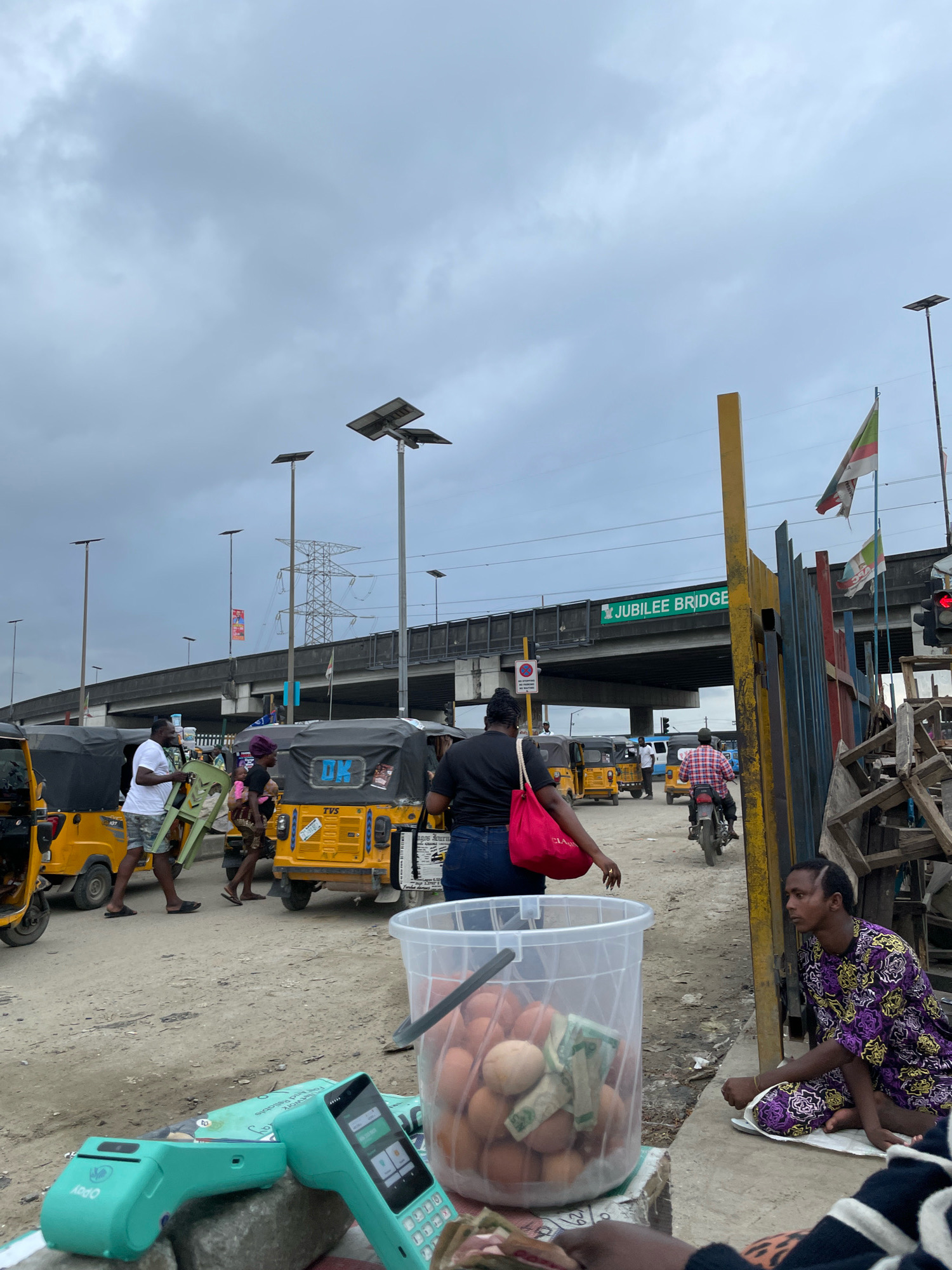 This image depicts a street scene in Ajah, Nigeria, under an overcast sky. The foreground features a clear plastic bucket filled with eggs and some currency notes, along with a teal-colored point-of-sale device and another card reader. To the right, a man wearing a purple and black patterned robe sits on the ground.
In the mid-ground, several yellow and black auto-rickshaws, known as "Keke NAPEPs," are parked or moving. A woman in a dark top and blue jeans, carrying a red bag, walks away from the viewer. Another woman holds a baby. A man in a white t-shirt and patterned shorts carries a green plastic chair. A man on a motorcycle wearing a red and black checkered shirt rides past.
The background shows an elevated road, identified by a green sign as "JUBILEE BRIDGE," with power lines and streetlights. Two flags, one with green, red, and white stripes and another with green and white, are visible. Construction materials are piled up to the right of the Jubilee Bridge sign. The ground is dirt and gravel.