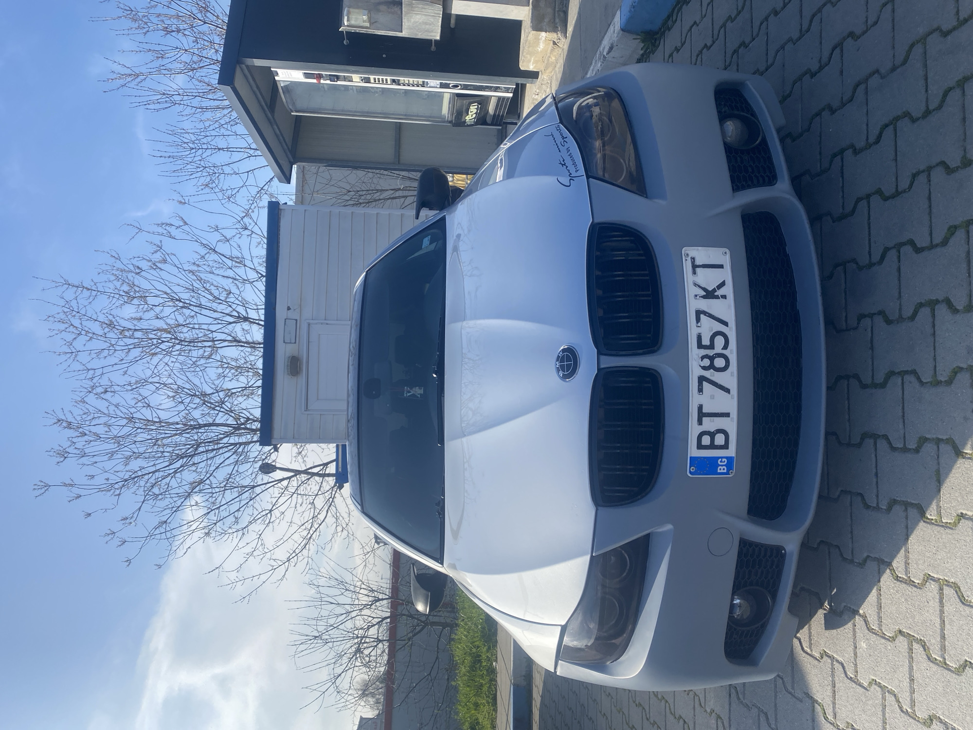 The image shows the front of a gray BMW car parked on a paved surface. The license plate reads "BT 7857 KT" and indicates that it's registered in Bulgaria. The car is parked near a small building or structure. In the background, there is a barren tree and a partial view of a cloudy sky. The location appears to be in an urban setting. While the image does not contain sufficient information to place the image in a precise location within Varna, the license plate suggests this photo was taken somewhere in Bulgaria.