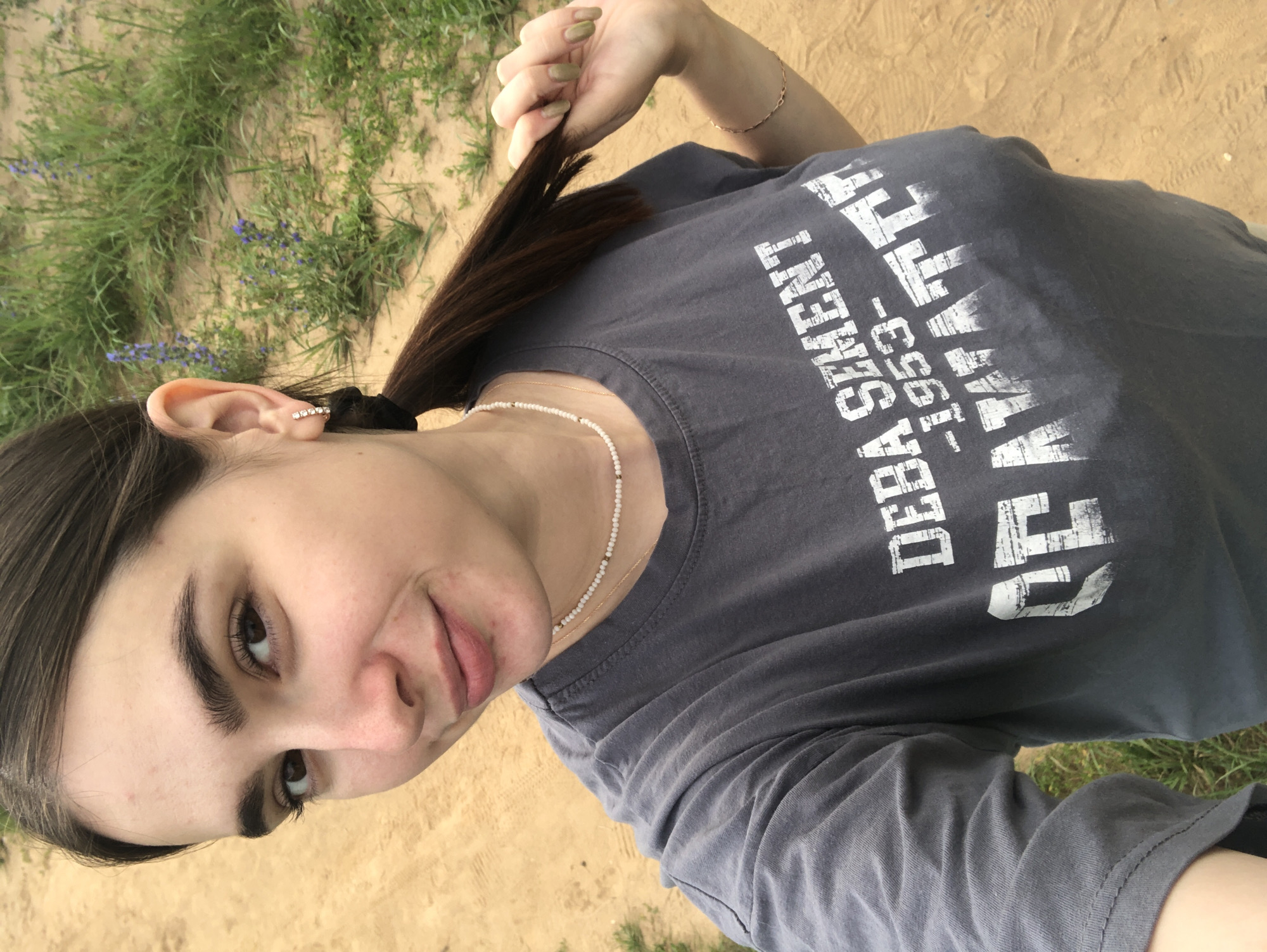 A young woman is taking a selfie outdoors in an area with dirt and sparse grass. She has long, dark brown hair pulled back in a ponytail and is wearing a grey t-shirt with white text. She is also wearing a white beaded necklace and a bracelet on her right wrist. Her right hand is holding a section of her ponytail. The background shows more grass and dirt, with some small purple flowers visible. The woman is smiling slightly. The image appears to have been taken in the city of Noviy, Russia.
