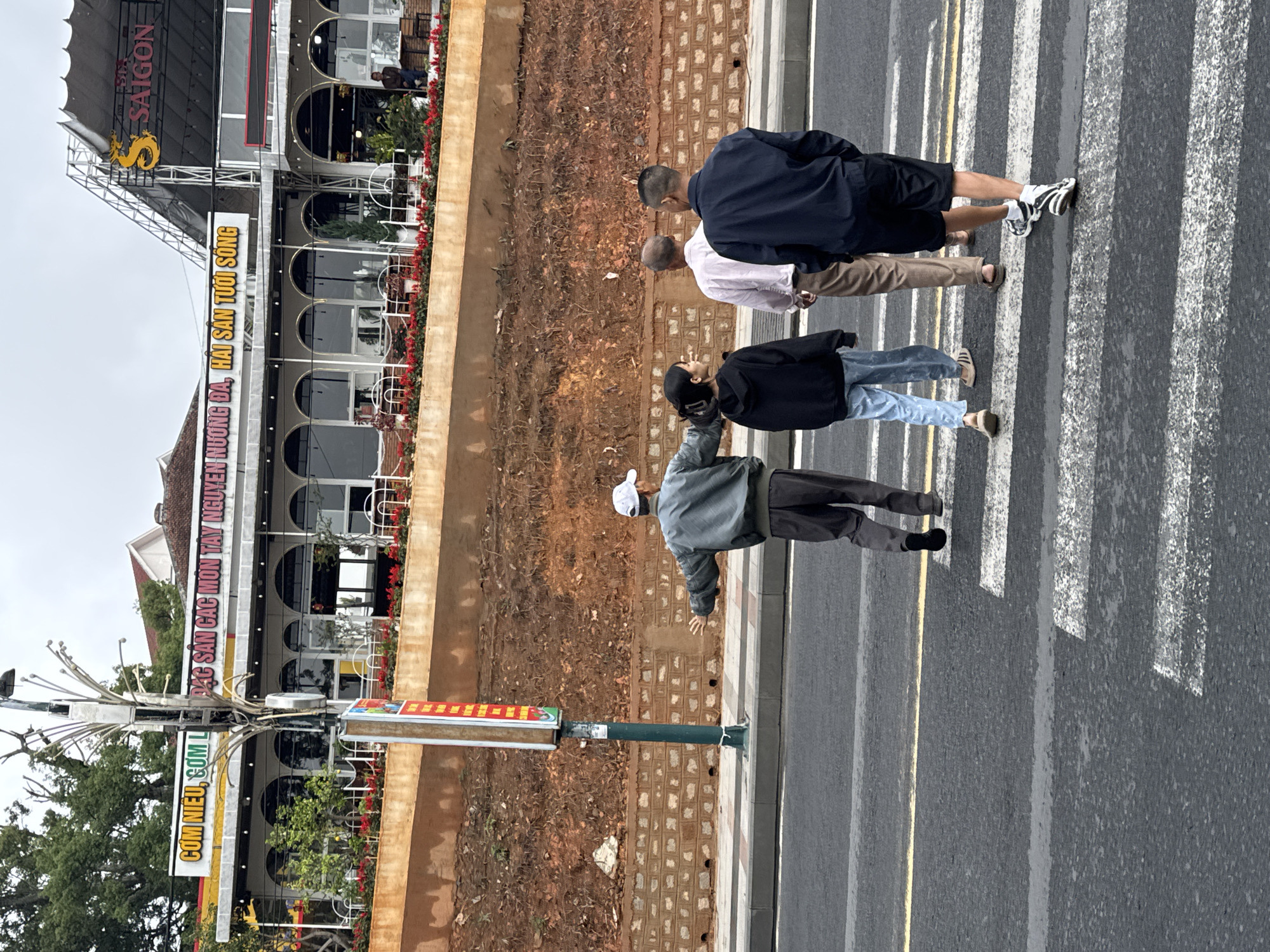 Four individuals are captured walking across a zebra crossing on a paved road, moving towards a row of buildings. The scene appears to be an urban street in Da Lat, Vietnam, under an overcast daytime sky, indicated by the soft, shadowless lighting.
From left to right on the crosswalk, the first person wears a white cap, a grey jacket, and dark pants, with an arm slightly extended. Next is a person in a black cap, black hoodie, and light blue jeans. Following them is an individual in a light-colored collared shirt, brown pants, and sandals. The last person on the right wears a dark blue jacket, black shorts, and black and white sneakers.
To the left of the road, an unpaved embankment with sparse vegetation separates the street from a multi-story building. This building features multiple arched windows and balconies adorned with red flowers. Prominent signs on the building display Vietnamese text. The top sign, accompanied by a golden dragon logo, reads "SAIGON". Below it, another sign states "YÊN NƯƠNG ĐÀ, HẢI SẢN TƯỚI SÔNG". Further down the street, on an adjacent building, another sign reads "CƠM NIÊU, CƠM ĐẶC SẢN CÁC MÓN TÂY NGUYÊN". A street light pole with a vertical, partially obscured sign stands by the roadside. The road itself has a clear zebra crossing and yellow dividing lines.