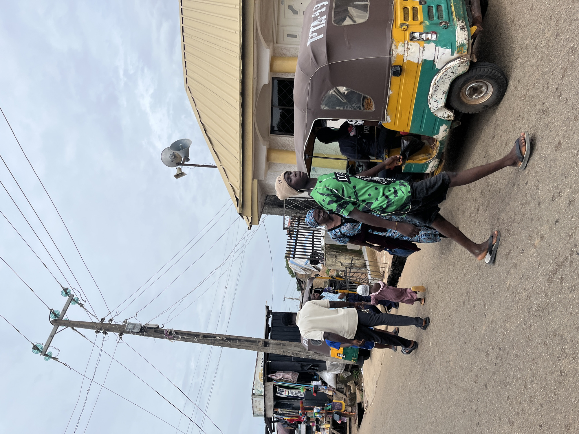 In Jos, Nigeria, on a cloudy day, a street scene unfolds with pedestrians and vehicles. A young man in a green and black t-shirt and black shorts walks towards the right, wearing a beige beanie and black flip-flops, with white earbuds in his ears. To his left, an older woman in a blue and black patterned dress and a blue headscarf walks away from the viewer. Behind them, a young child in a pink outfit and white cap walks towards the left, holding hands with an adult whose legs are visible. Further left, a man in a white t-shirt and black pants walks away from the viewer, followed by a person in a blue shirt.
A yellow, green, and white auto-rickshaw, also known as a Keke Napep, is parked to the right of the frame, facing left. The front of the auto-rickshaw is brown, and it displays the lettering "PTA-49" on its side. A dark-skinned person is seated in the driver's seat, and another person is visible behind them.
On the left side of the street, a makeshift stall is set up with various items displayed for sale, including bags and other merchandise. A tall wooden utility pole with multiple wires and insulators stands prominently on the left side of the street, with a meter box attached to it. In the background, a building with a yellow corrugated roof and cream-colored walls is visible. Another building with a black exterior and a metal roof is situated behind the stall. The street itself is paved with asphalt.