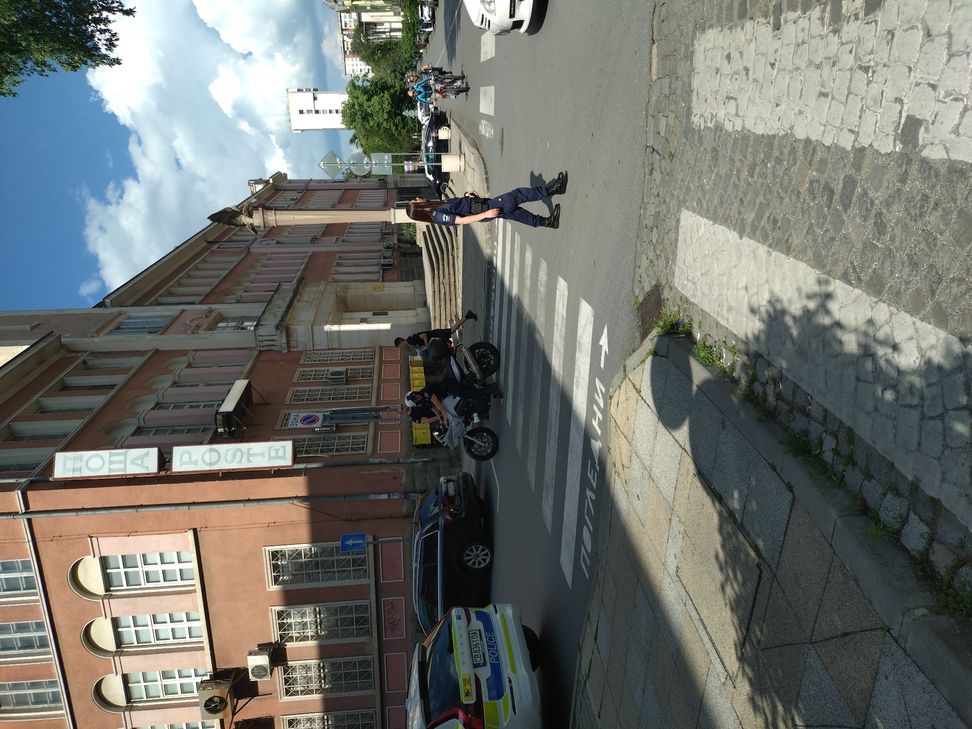 The image shows a street scene in Stara Zagora, Bulgaria. A police officer is standing in a crosswalk. A police car and a police motorcycle are parked on the street. A building with a sign that reads "ПОЩА POSTE" is visible. There are other cars and people in the background. The sky is blue with some clouds.